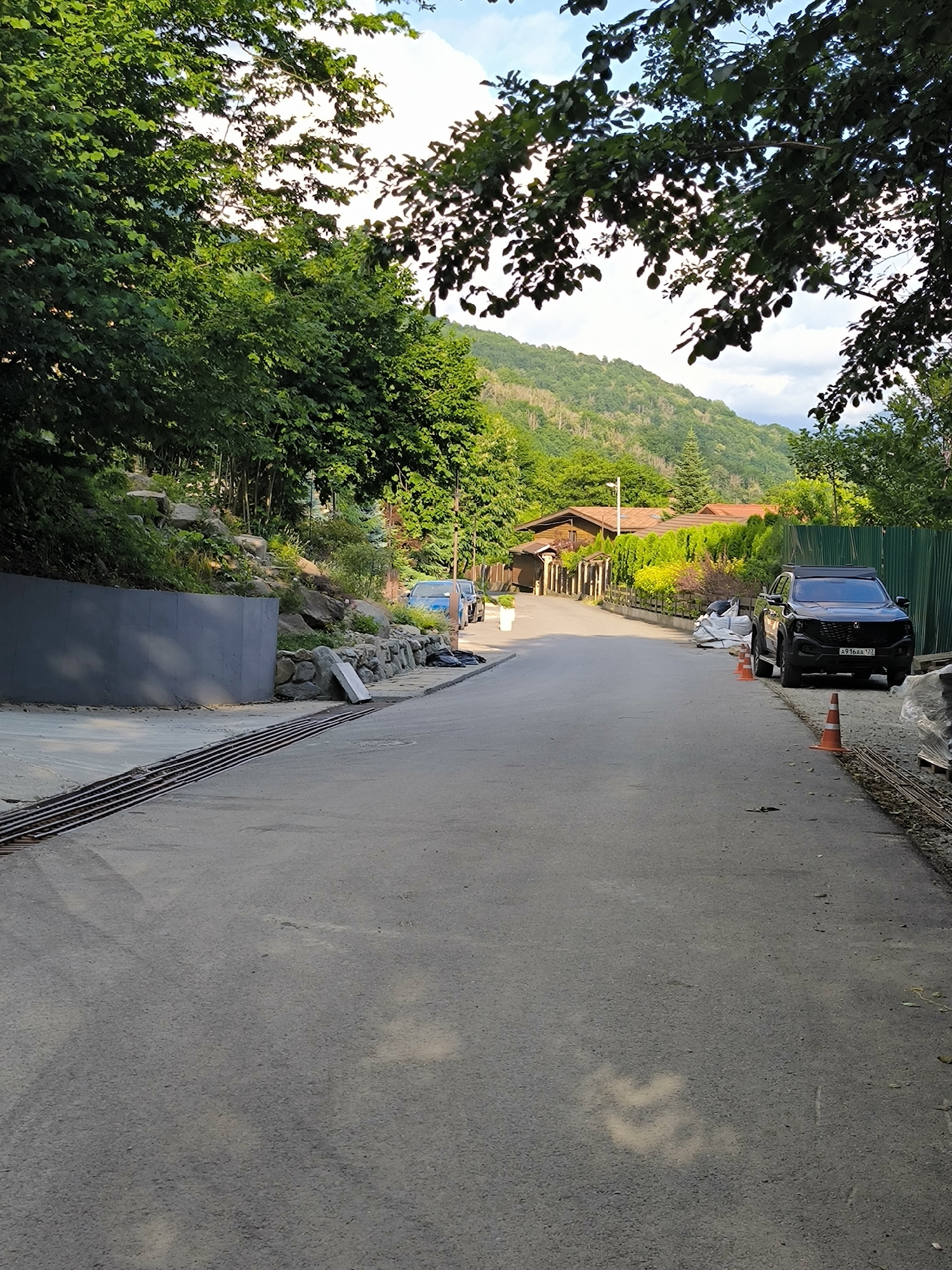 The image depicts a street in the city of Krasnaya Polyana, Russia, likely a residential area given the presence of houses and trees. The street is paved and appears to be a two-lane road. On the left side of the road, there is a grey concrete wall and a rock retaining wall with vegetation growing on it. A metal grate is visible on the pavement near the wall. On the right side of the road, there is a black SUV parked, and construction materials, including bags and orange traffic cones, are present. The street leads towards a residential area with houses that have reddish-brown roofs, surrounded by greenery and trees. In the background, a lush green hillside is visible, suggesting a mountainous or hilly terrain characteristic of Krasnaya Polyana. The sky is partly cloudy. No people are visible in the image. The scene appears to be a quiet, somewhat rural street with some ongoing construction or road maintenance.