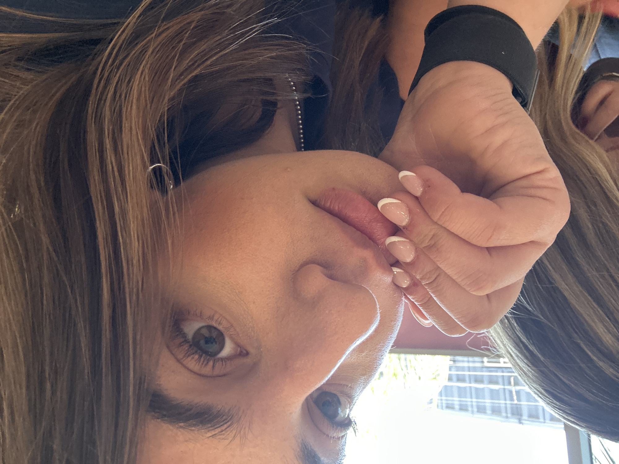 The image displays a close-up, slightly upward-angled perspective of a young female individual. She has light to medium skin, long brown hair with lighter highlights, and dark brown eyes looking directly forward. Her left hand is raised, with fingers touching her chin and lower lip. The fingernails of her left hand exhibit a French manicure style with a light pink base and white tips. A black wristband is present on her left wrist. She wears a small silver hoop earring in her right ear and a silver beaded chain necklace, partially visible below her chin. To the far left, partially obscured, is another individual's head and upper back, showing blonde-highlighted hair.
The background consists of an interior space with a large window or glass panel. Through this window, a modern multi-story building with a distinctive grid-like pattern of numerous windows is visible under bright daylight conditions. A reddish-pink surface or wall section is visible to the right of the window frame.
The image does not contain any specific identifiable landmarks, street signs, or unique architectural features that would allow for the determination of a precise location within the city of Puebla, Mexico. The visible background building is generic in nature.