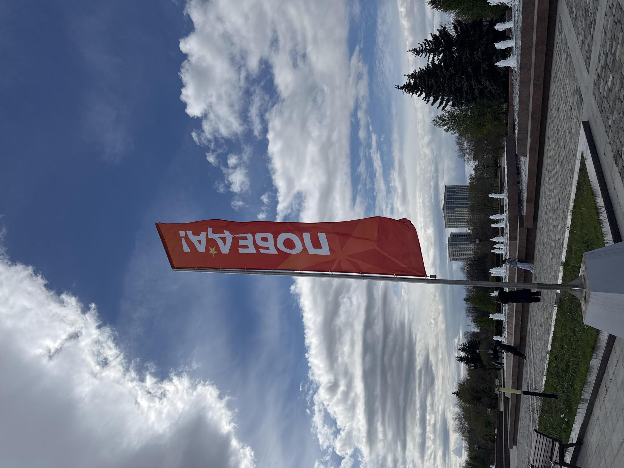The image shows a red flag with the word "ПОБЕДА!" (Victory!) in white Cyrillic letters, against a blue sky with white clouds. The flag is mounted on a pole, and the bottom of the pole is visible.
In the background, there is a park with trees, fountains, and buildings. The location is most likely Victory Park in Moscow, Russia, based on the flag and the style of the park architecture.
There are people in the background near the fountains. A bench is visible in the lower right corner. The pathway is paved with stone. The weather appears sunny.