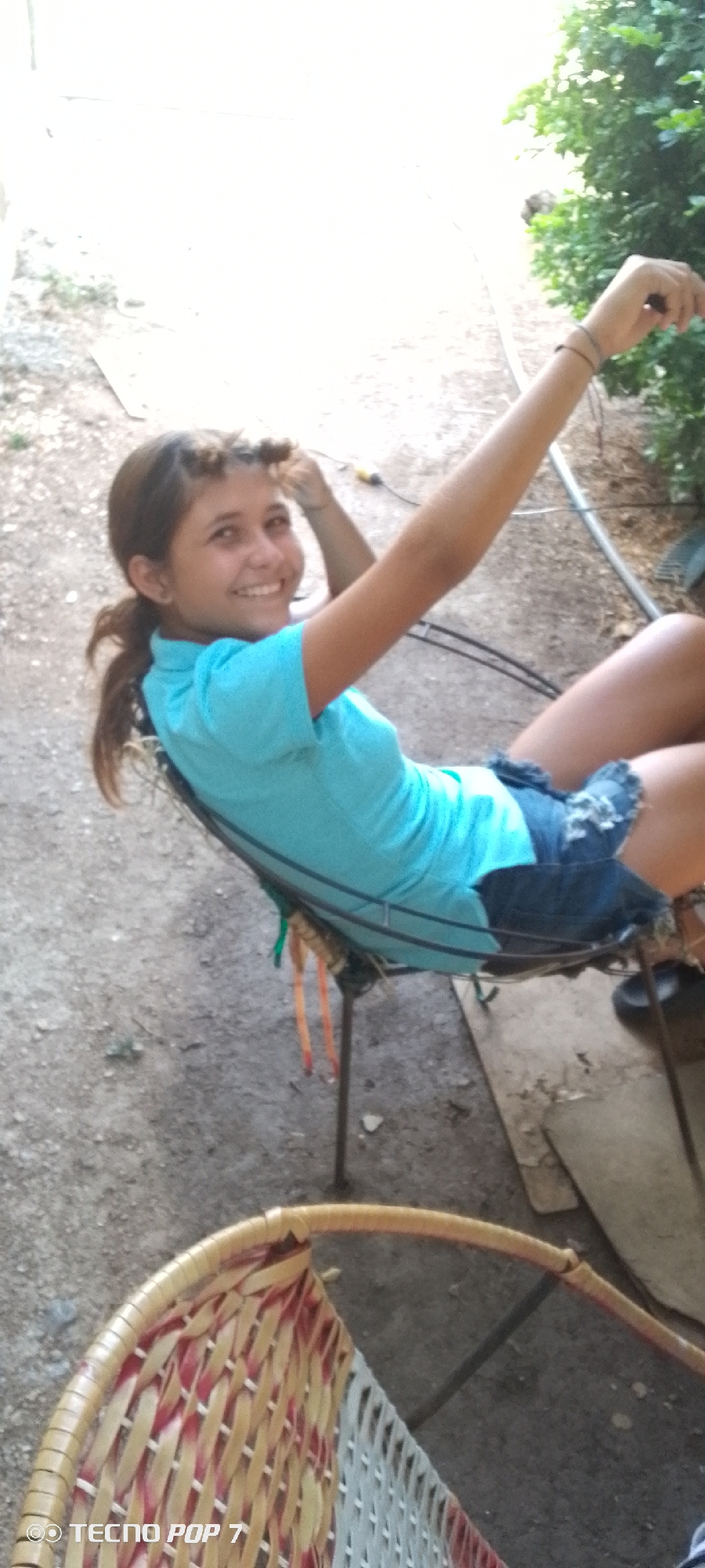 An outdoor portrait captured in a slightly tilted orientation features a young girl, likely a pre-teen or teenager, sitting in a metal-framed chair. She is smiling broadly and looking directly at the camera, conveying a joyful or content emotion. She wears a light blue short-sleeved t-shirt and denim shorts. Her dark hair is pulled back, with some strands appearing to be styled or bunched near her forehead, and a ponytail is visible behind her left shoulder. Her right arm is raised and bent at the elbow, with her hand near the upper right edge of the frame, while her left hand is closer to her head.
The setting appears to be an unpaved outdoor area, possibly a yard, with visible dirt and gravel on the ground. Lush green foliage and bushes are present in the background on the right side. The lighting is bright, indicating it is daytime, with some areas in the background appearing overexposed due to strong sunlight. A cable or cord is visible hanging behind the girl's head and right arm. The chair she is seated in has a simple metal frame, and several colorful straps or pieces of fabric hang from its side. Partially visible in the bottom left foreground is another chair, featuring a woven rattan or wicker design with patterns of natural, red, and white fibers.
The watermark "TECNO POP 7" is visible in the bottom left corner of the image, indicating the device used to capture the photo. The overall scene suggests a casual moment outdoors in Altagracia, Venezuela.