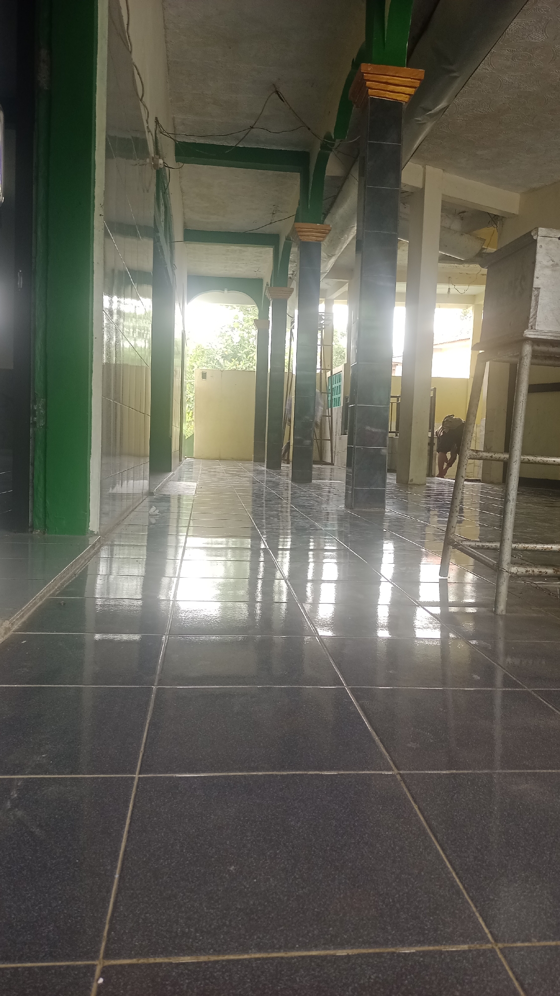 The image depicts a wide hallway or corridor with a tiled floor that is highly reflective due to a recent cleaning or natural moisture. The hallway is lined with dark, square tiled pillars, some of which are painted green and accented with yellow at the top. The ceiling appears to be concrete with exposed beams, some of which are also painted green. Natural light streams in from an open area at the end of the hallway, illuminating the reflections on the floor and highlighting the details of the pillars.
In the background, a person can be seen walking away from the camera, partially obscured by a pillar and some outdoor greenery. The setting appears to be an open-air structure, possibly a mosque or a communal building, given the architectural style and the presence of the pillars. The tiled walls on the left suggest a covered walkway or an adjacent room. The overall impression is one of a clean and well-maintained public space in Karyamandala, Indonesia, characterized by its reflective surfaces and functional design.