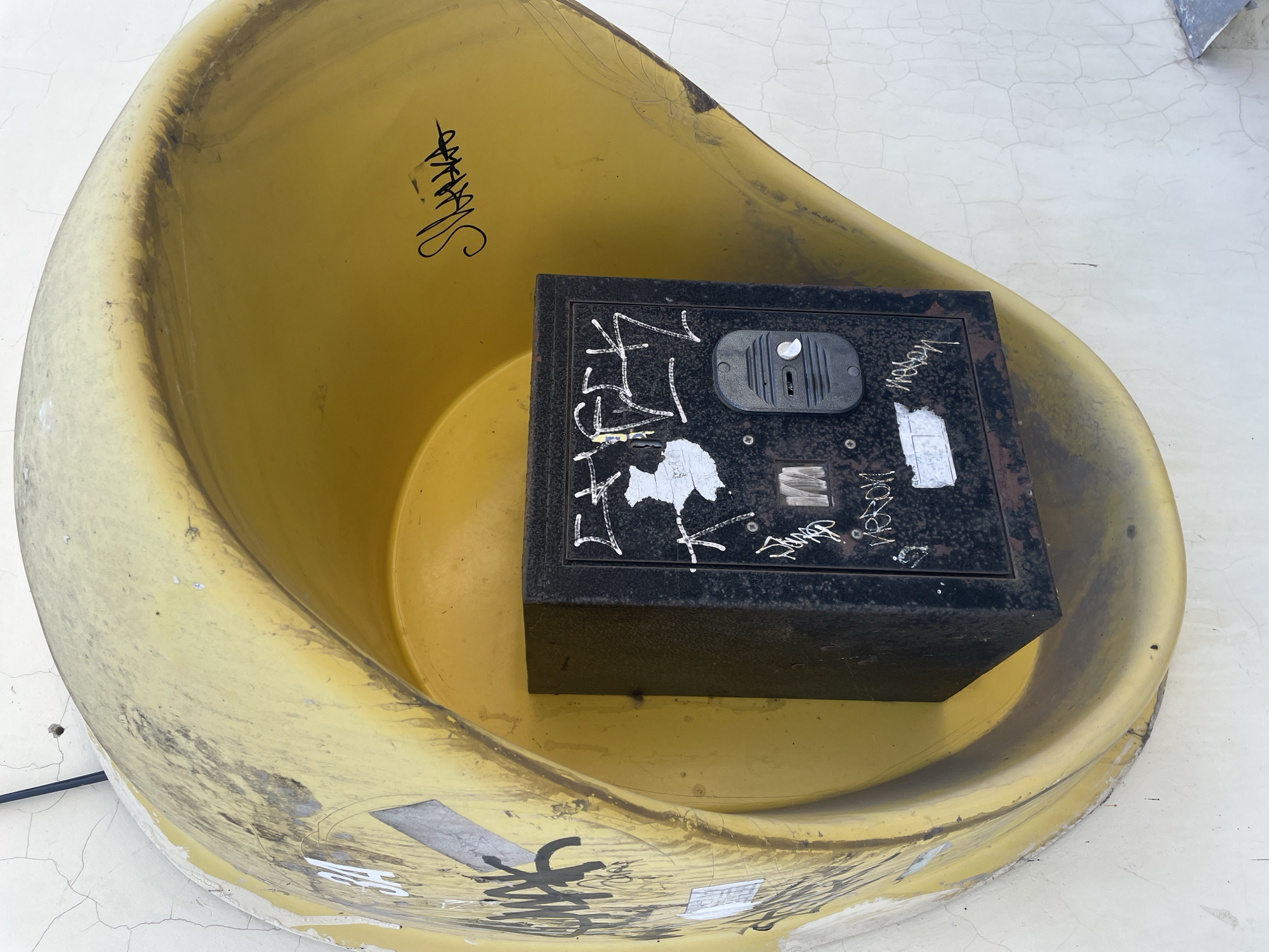 The image shows a close-up, eye-level view of a black metal safe placed inside a weathered, yellow, shell-like structure. The safe is square and appears to be made of rusted metal. It has a gray electronic lock or keypad on its front. White graffiti, some of which is illegible, is scrawled across the safe's surface and also covers parts of the yellow structure. The yellow structure is mounted on a light-colored, cracked wall. The lighting suggests it is daytime. There are no people or animals visible in the image. The overall scene appears to be outdoors, possibly in an urban environment, given the graffiti and the utilitarian nature of the objects. The specific location is not discernible from the image itself.