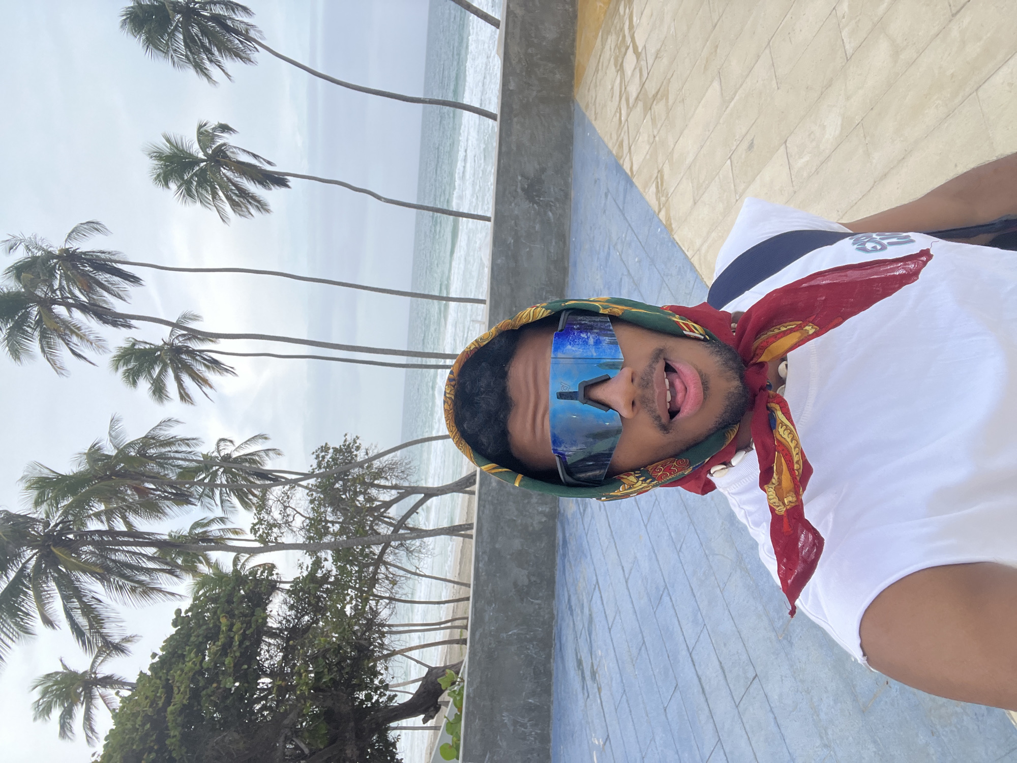 A selfie-style, rotated eye-level shot taken outdoors on a cloudy day. The subject is a Black male wearing sunglasses, a scarf, and a white T-shirt. He is sticking his tongue out slightly. Behind him is a beach scene, which includes palm trees and the ocean. The location is consistent with Playa El Agua, Venezuela. There is a low wall in the immediate background, separating the beach from a paved area. The pavement is made up of a light yellow and blue tiled design.