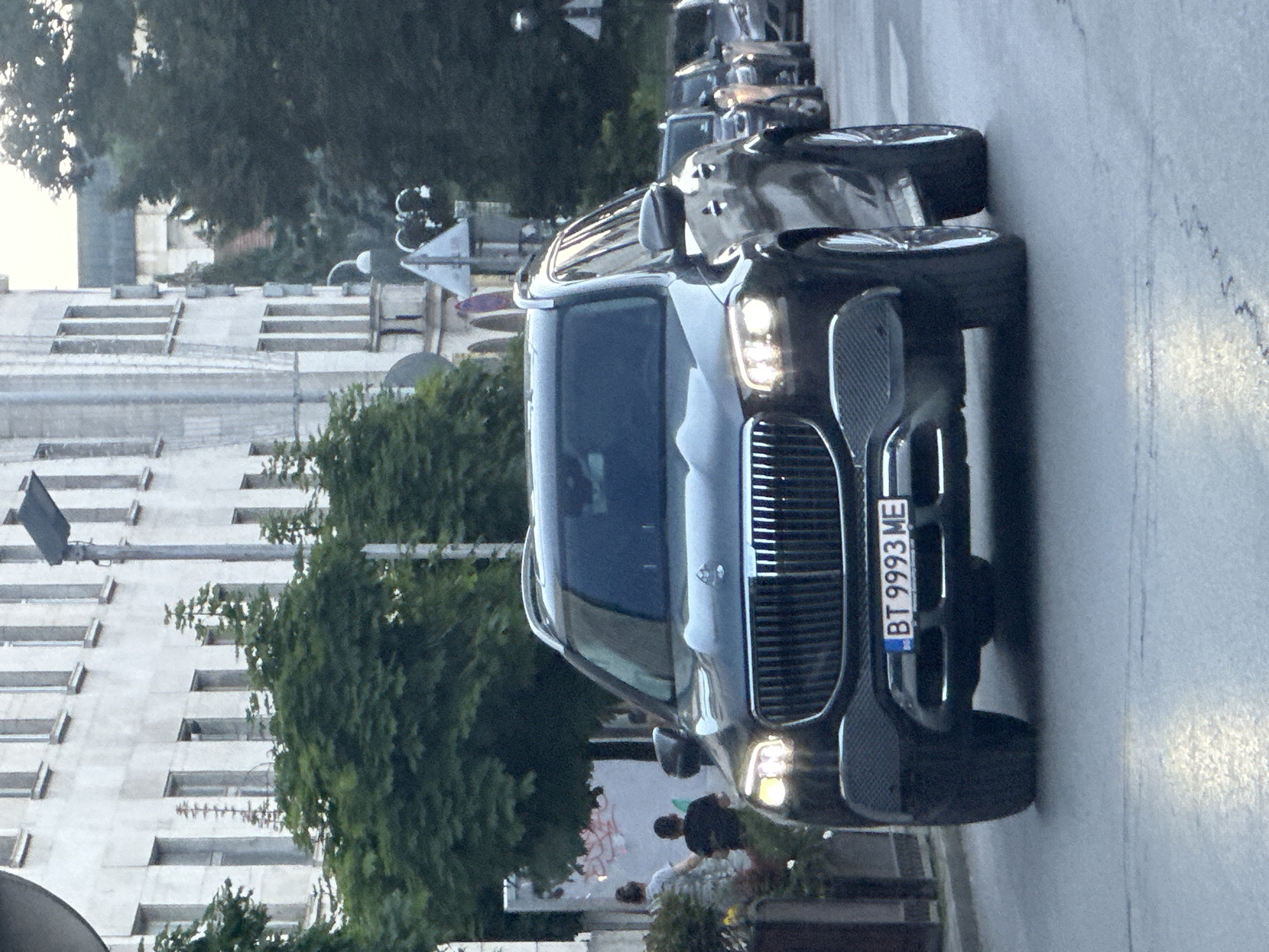 The image shows a black car, possibly a Mercedes-Benz Maybach GLS, parked on a street in Veliko Tarnovo, Bulgaria. The license plate reads "BT 9993 ME". In the background are buildings, trees, and other vehicles. People are present near the car. The street appears to be a typical city street.