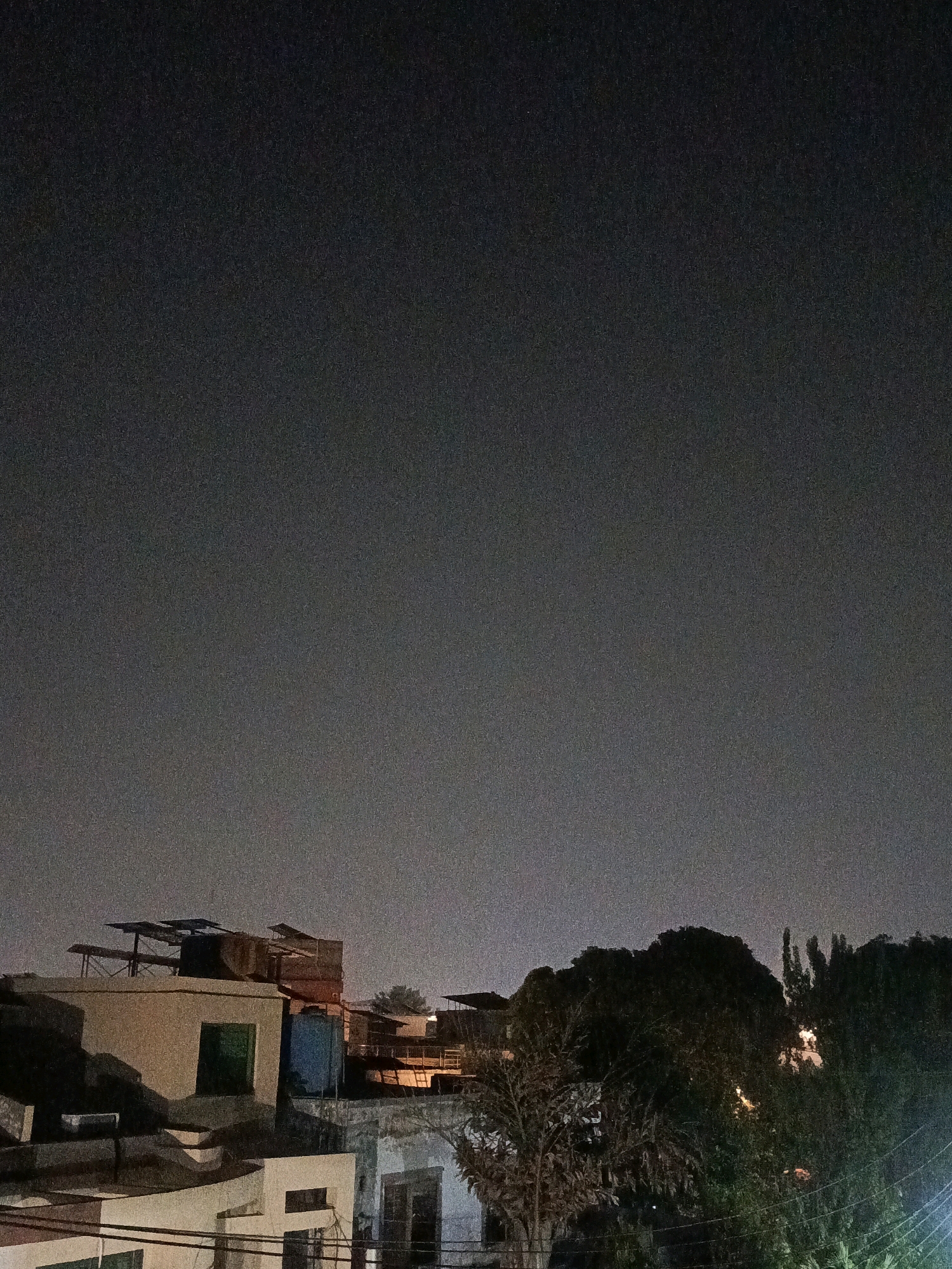 The image depicts a nighttime scene in the city of Rasoolpur, Pakistan, focusing on the rooftops of buildings under a dark, cloudy sky. In the foreground, the tops of several buildings are visible, characterized by flat roofs, varying architectural styles, and some with visible water tanks. One prominent structure has a light-colored facade with a green-colored window and a blue water tank on its roof. Further back, a dense cluster of trees obscures parts of the buildings and skyline. The overall ambiance is dark and grainy, suggesting low-light photography. There are no people visible in the image, and no specific actions or interactions are occurring. The environment appears to be an urban or semi-urban residential area.