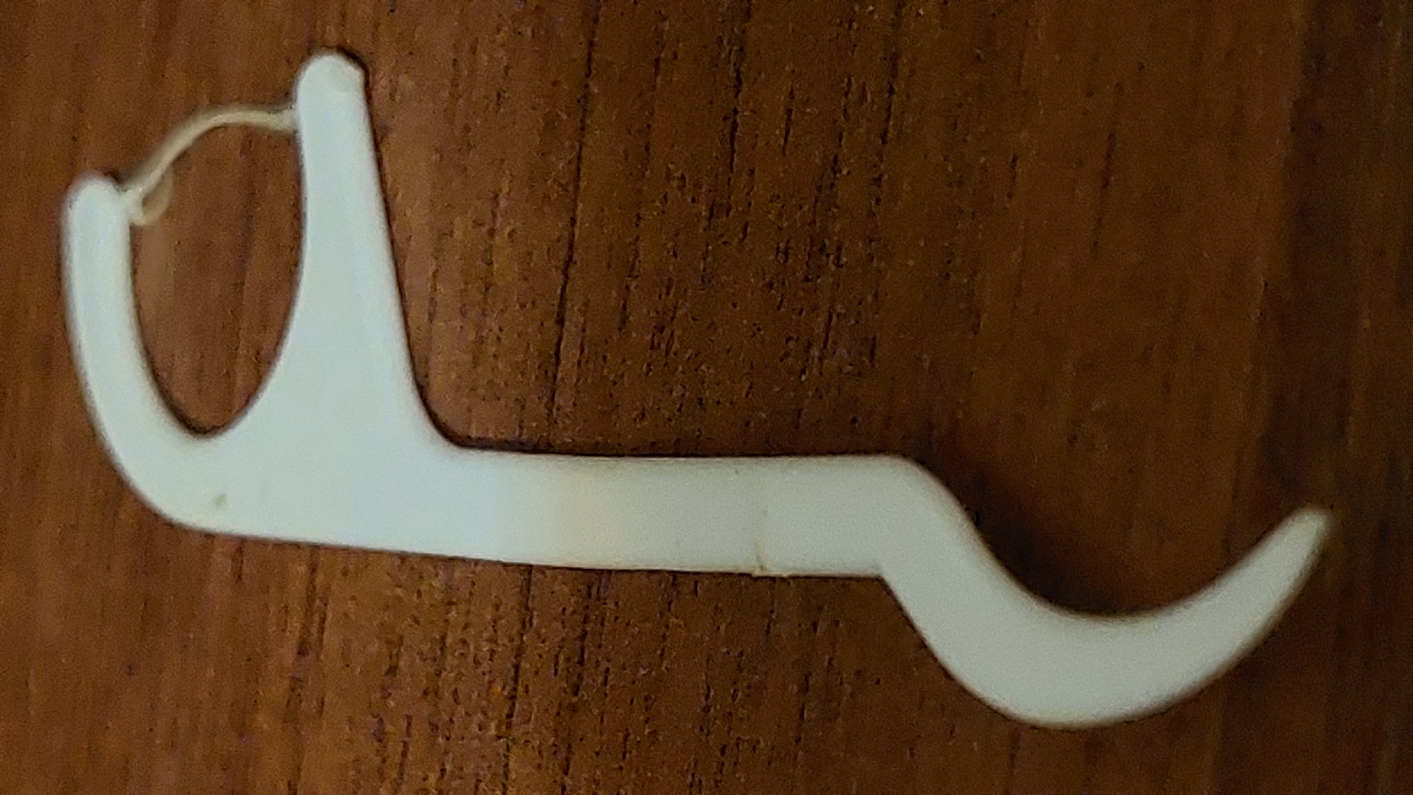 A white dental floss pick with a curved handle and a looped end holding a strand of floss rests on a dark brown, wood-grained surface. The floss appears slightly yellowed or soiled. The object is positioned diagonally across the frame, with the handle curving towards the bottom left. The lighting is overhead, casting soft shadows and highlighting the texture of the wood. There are no people present. The location is not discernible from the image alone.