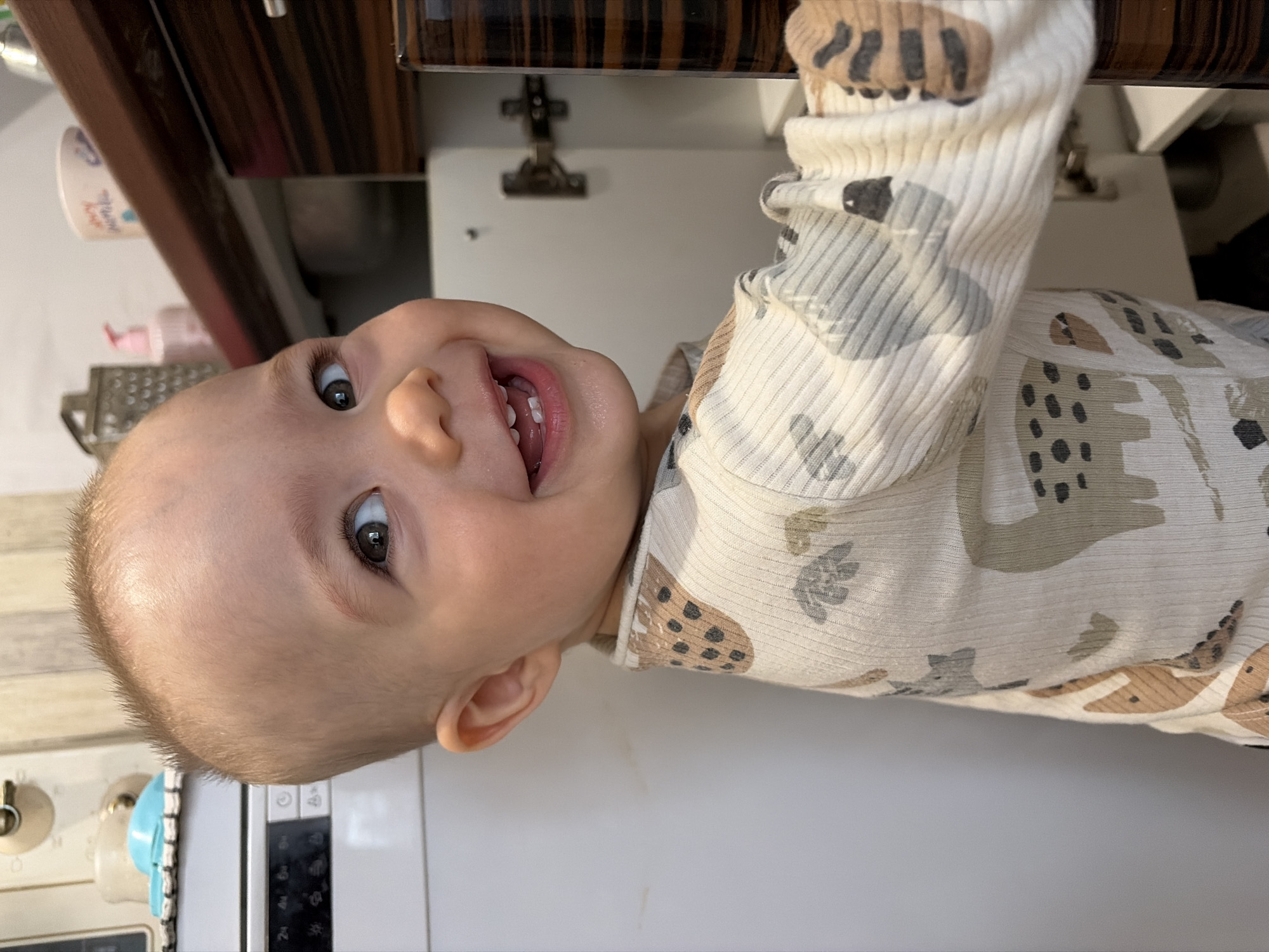 A baby wearing a patterned, long-sleeved shirt is lying on its back in a kitchen, with its head near a white microwave and kitchen cabinets. A grater and bottles of soap are visible on the countertop. It cannot be determined if the location is the city of Chaikovsky, Russia.