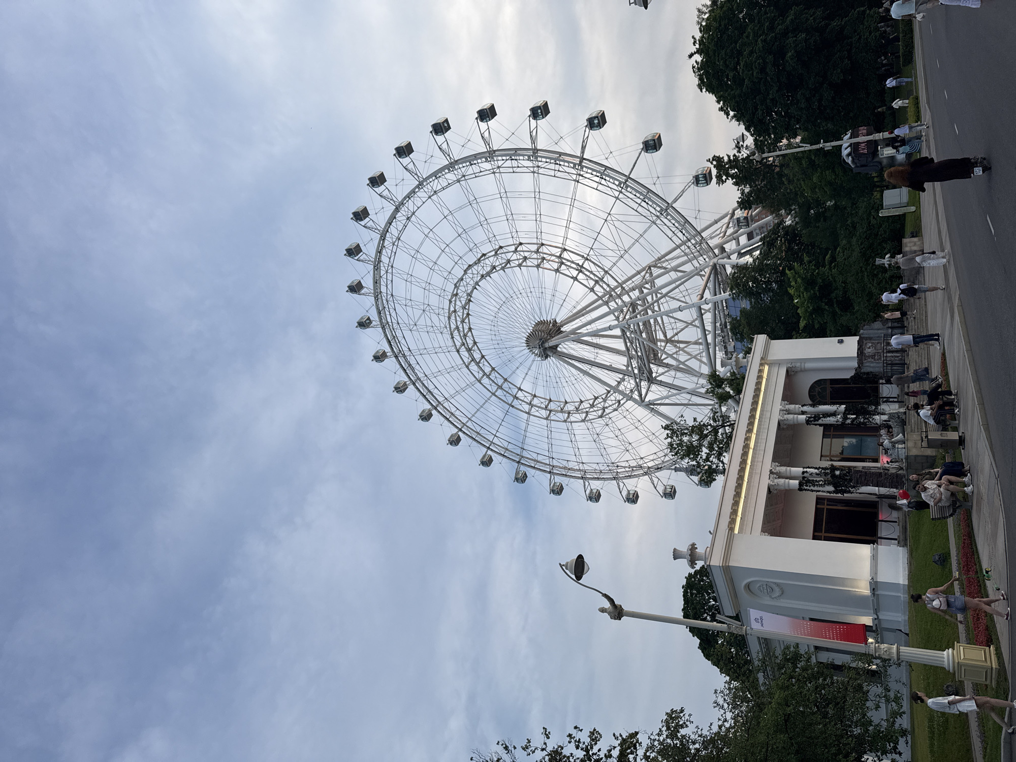 This is a wide-angle, eye-level, outdoor shot of the Ferris wheel at VDNKh Park in Moscow, Russia. The massive white Ferris wheel dominates the upper right portion of the frame, with its intricate metal structure and numerous enclosed cabins visible against a cloudy sky. In the foreground, a white, neoclassical building with columns and arched windows stands to the left, partially obscured by trees. A lamppost with an ornate base is positioned in front of the building. People are scattered throughout the scene, walking along a paved pathway in front of the building, sitting on benches, and one person is riding a scooter on the road. The surrounding area is green with trees and grass.