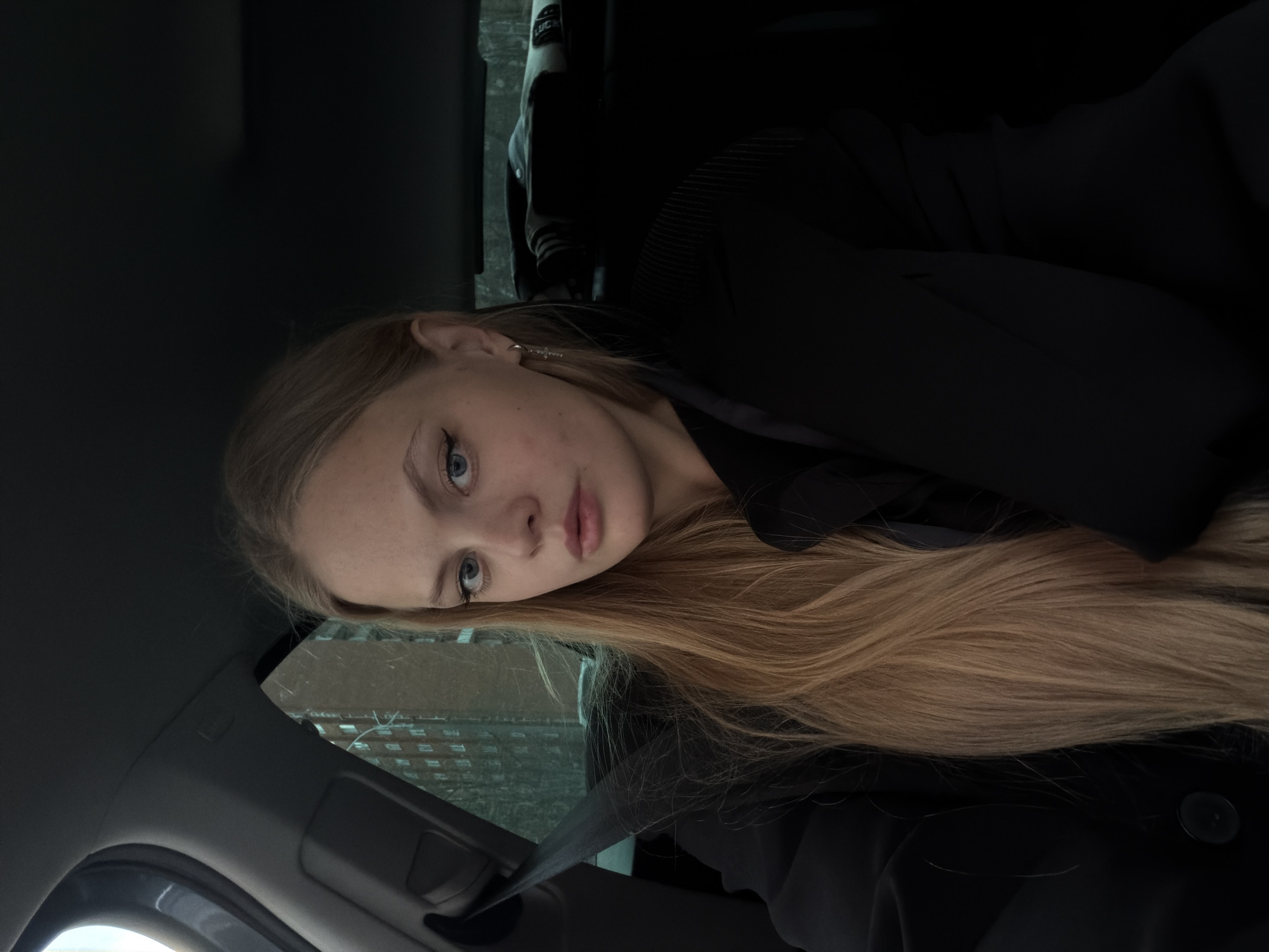 A young woman with long, straight blonde hair and blue eyes is pictured inside what appears to be a car, possibly in Izhevsk, Russia. Her head is tilted, and she looks directly towards the camera with a calm and slightly contemplative expression. She has light-toned skin and visible eyeliner. She is wearing dark clothing, possibly a black jacket or coat, and a car seatbelt is fastened across her chest, suggesting she is either a passenger or a driver.
The car's interior is visible, including the dark headliner, parts of the front overhead console, and the seatbelt mechanism. Through the car window in the background, a large, multi-story building with numerous windows is visible, indicating an urban or residential environment. The lighting appears to be natural daylight, potentially overcast. In the darker upper background, behind her head, a partially visible light-colored logo spells "LUCK" on what seems to be another piece of clothing or an item. The overall scene is quiet and captures a momentary gaze from within a vehicle.