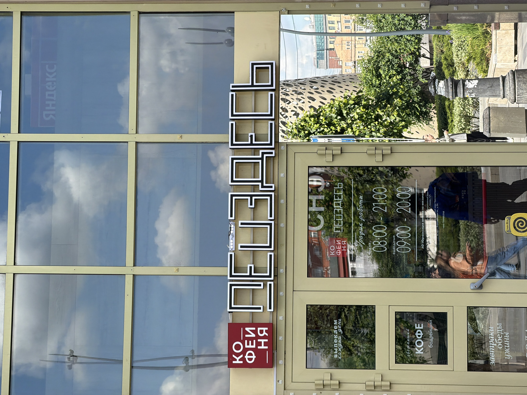 The image captures the exterior of a commercial establishment, likely a bakery and coffee shop, on a bright, sunny day in Moscow, Russia. The building features a facade of large glass windows and doors set within light beige or pale yellow frames.
The prominent signage includes a large white sign with black outlines spelling "ПЕКАРНЯ" (Bakery) in Cyrillic. Below it, a smaller red sign displays "КОФЕЙНЯ" (Coffee Shop).
Numerous reflections in the glass reveal the surrounding urban environment. White clouds dot a clear blue sky, while distant buildings, lush green trees, bushes, and a dark street lamp are visible. Reflections of people, including one wearing a light-colored top and jeans, suggest active pedestrian traffic outside.
Detailed text on the door provides further information about the business:
- It reiterates the name "ПЕКАРНЯ. КОФЕЙНЯ."
- "График работы:" (Working hours:) are specified as "будни 08:00 - 21:00" (weekdays 08:00 - 21:00) and "выходные 09:00 - 20:00" (weekends 09:00 - 20:00).
- Services advertised include "выпекаем каждый день" (we bake every day), "свежайший КОФЕ бодрейший" (freshest coffee most invigorating), and "завтраки обеды ужины" (breakfasts lunches dinners).
- Corporate details "АО «Организация питания на вокзалах»" (JSC "Catering Organization at Railway Stations") and "ИНН 7709883768," along with "округ Красносел" (Krasnoselsky District), indicate a likely location near a railway station in Moscow.
- A "Яндекс" (Yandex) logo is reflected in an upper window, and a "Яндекс Еда" (Yandex Food) logo is visible on the lower part of the door, suggesting delivery services.
The image is notable for its vertical orientation, capturing the scene as if the camera was held sideways.