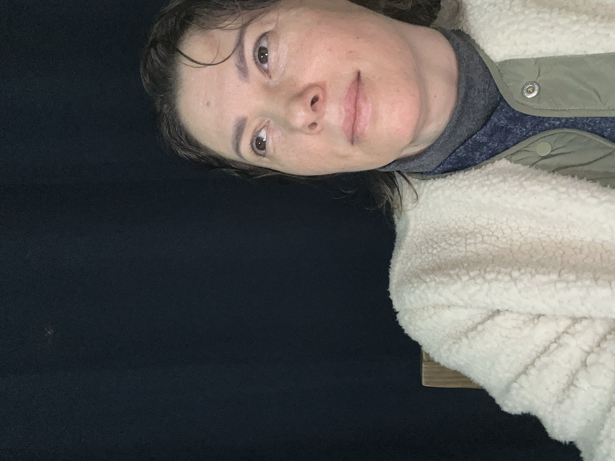 A medium shot captures a woman looking upwards, her face angled towards the top right corner of the frame. She has medium-toned skin, brown hair, and brown eyes. She is wearing a cream-colored fuzzy jacket over a dark grey turtleneck and a blue patterned sweater. Her left shoulder is partially obscured by a wooden chair back. The background is a dark, solid color, likely a curtain or wall. There is a small, faint speck of light visible in the upper left quadrant of the image. The setting appears to be indoors. The location in Buenos Aires, Argentina, is not specified.