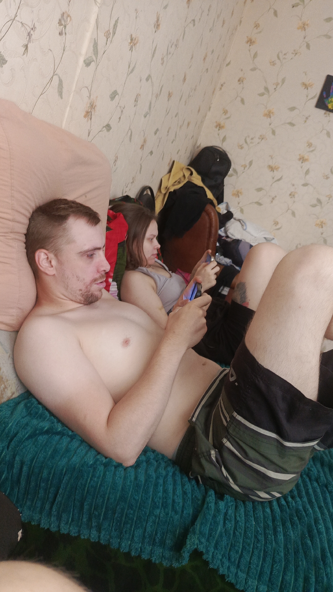 A man and a woman are lying down, possibly on a bed, facing away from each other. The man is shirtless, wearing dark shorts with green stripes. He is looking to his left with a neutral expression, his head resting on a tan pillow. The woman is wearing a grey tank top and dark shorts. She is holding a smartphone in her right hand and looking down at it. Her left hand is also on the phone. Both are lying on a teal, textured blanket. Behind them, there is a pile of clothes and a black backpack against a wall with floral wallpaper. The scene appears to be indoors, with artificial lighting. There is no visible text in the image.