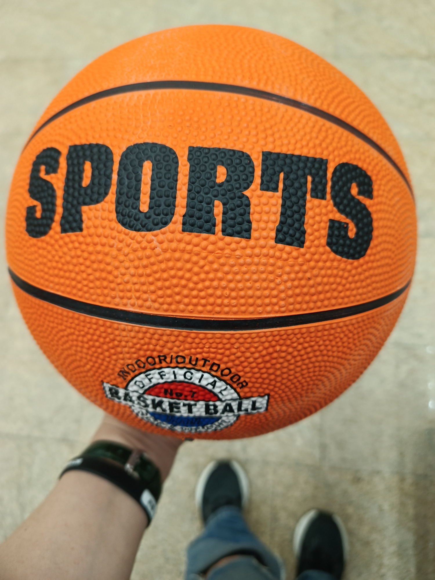 A person holds an orange basketball in their hand. The basketball has the word "SPORTS" printed in large, black letters on the upper half, with two black lines wrapping around the circumference of the ball. The lower half of the basketball has a circular logo that reads "INDOOR/OUTDOOR OFFICIAL BASKETBALL No.7." The person is wearing a black fitness tracker on their wrist. Below the hand holding the basketball, the person's feet wearing black shoes with white trim and light blue jeans are visible. The background appears to be a light-colored tiled floor. There is no indication of the location being in Moscow, Russia.
