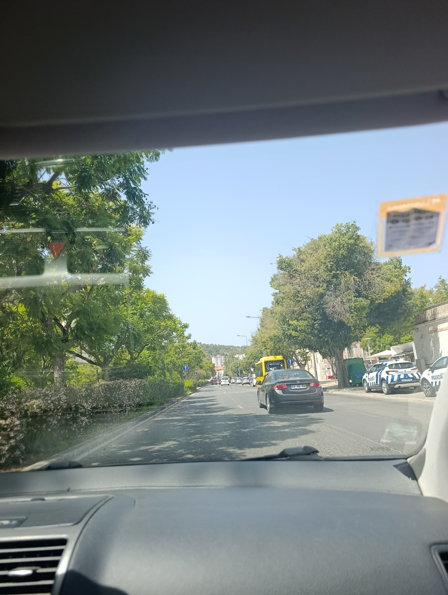 The image is taken from inside a vehicle, looking forward through the windshield. The scene depicts a street in Setúbal, Portugal, on a sunny day. A row of trees lines the left side of the road, with manicured bushes in the foreground. On the right side of the road, there are several police vehicles, identified by the word "POLICIA" on their sides. One police car is white with blue and black accents, and another appears to be similar. A yellow bus is visible further down the road, as well as other vehicles and buildings in the distance. The sky is clear and blue. The interior of the vehicle shows a portion of the dashboard and windshield frame.