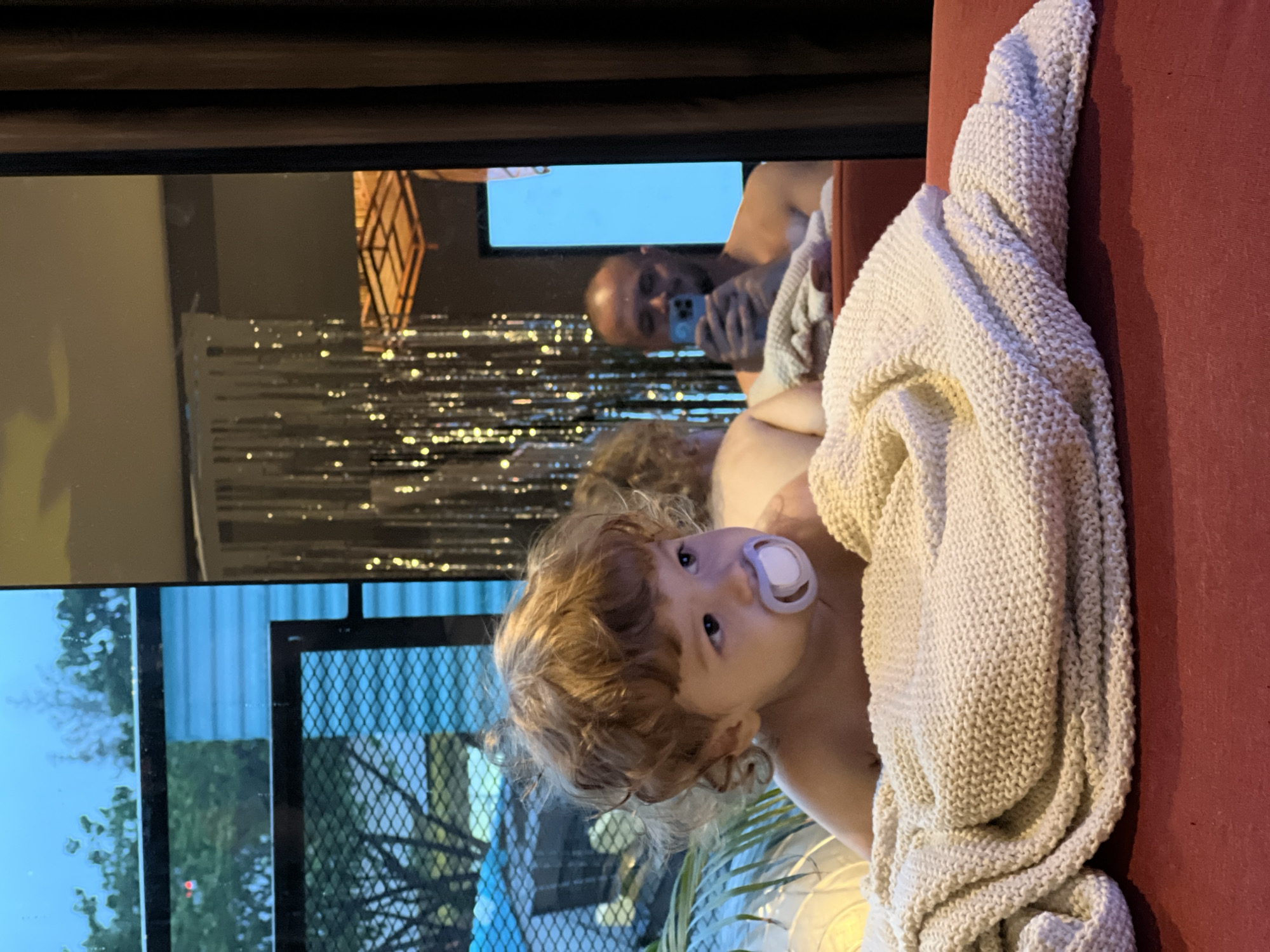 A toddler with curly light brown hair and a pacifier in their mouth is wrapped in a light beige knitted blanket. The toddler is positioned in the foreground, with their head and upper body visible. Behind the toddler, a man is reflected in a mirror, holding a blue smartphone and taking a selfie. The man is bald and appears to be shirtless. In the background, through a window or glass, trees and a blue-tinted building are visible. Decorative silver fringe hangs in the mid-ground of the mirror reflection. The scene appears to be indoors, with a dark curtain visible on the right side of the frame.