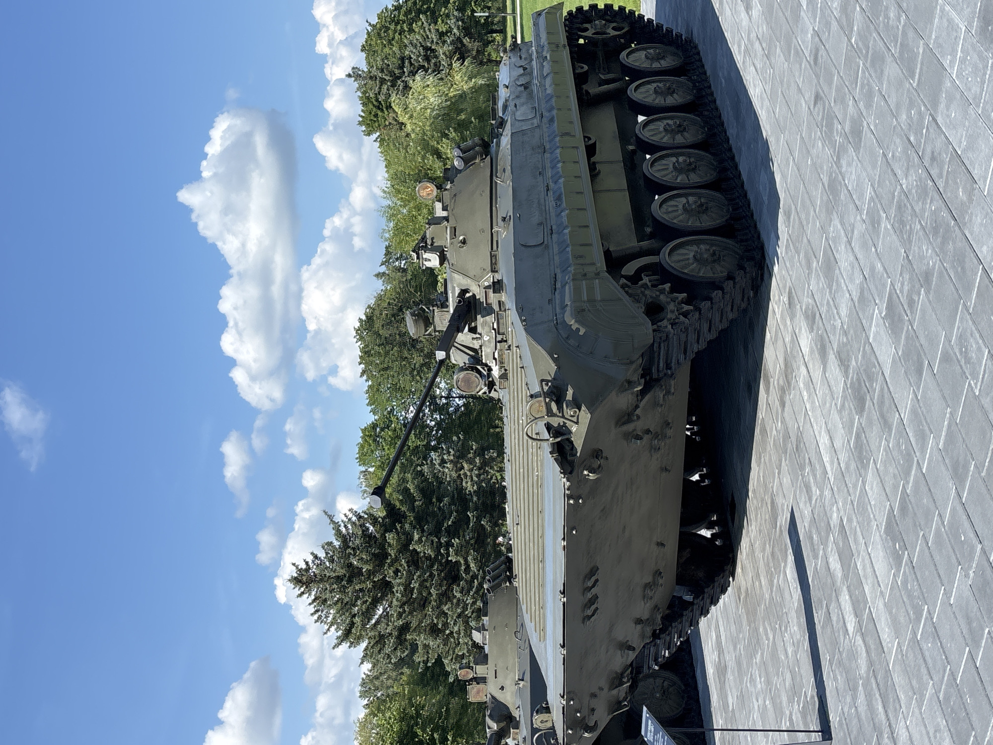 A weathered, olive-drab military armored personnel carrier sits on a paved courtyard in Ozyaritska-Slabadski rural council, Belarus. The vehicle, possibly a BMP-1, is stationary and displayed outdoors, likely as a memorial or exhibit. It is positioned at an angle, with its front facing slightly towards the viewer's left. The APC's tracks are resting on a light gray, rectangular paver surface. Behind the APC, a lush green park setting unfolds, featuring mature trees and a bright blue sky dotted with fluffy white clouds. The sun is casting shadows on the ground, indicating daytime. No individuals are visible in the frame.
