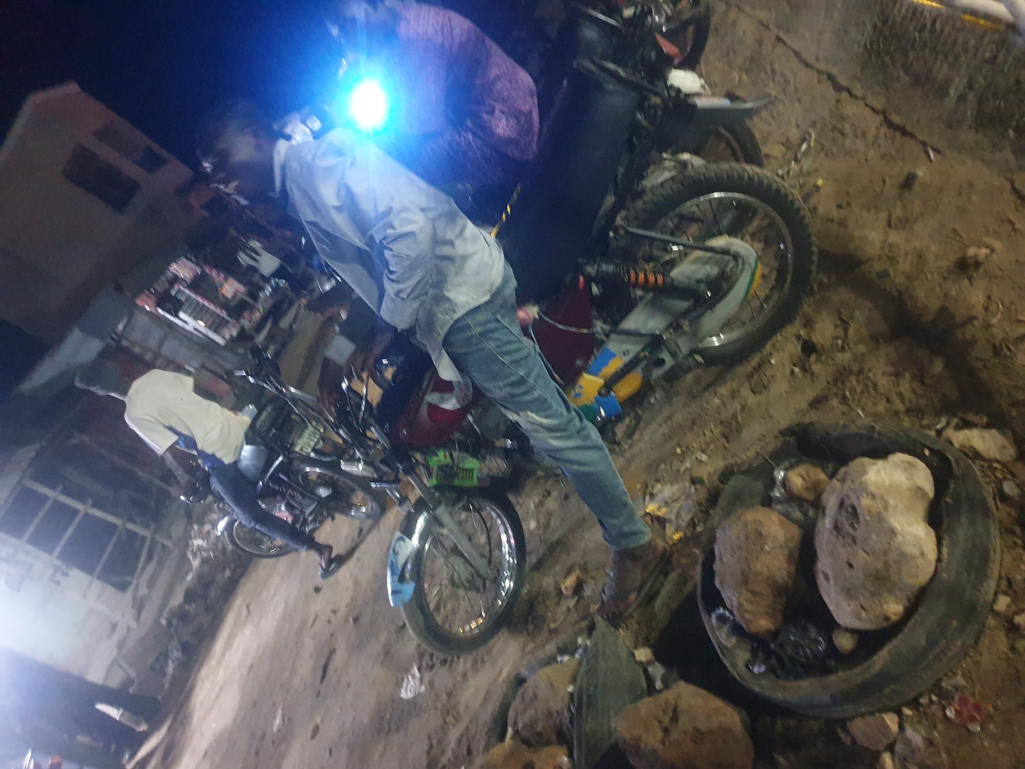 The image depicts an outdoor scene in what appears to be the city of Zango-ndaji, Nigeria, based on the surroundings. At least two motorcycles are visible, with one being larger and more prominent. A person wearing a light-colored shirt and jeans is standing next to the larger motorcycle. The environment is rugged, with dirt and debris visible. A container filled with rocks is in the foreground. Building structures are present in the background, possibly indicating an urban or semi-urban setting. A light source shines brightly, possibly from a motorcycle headlight.