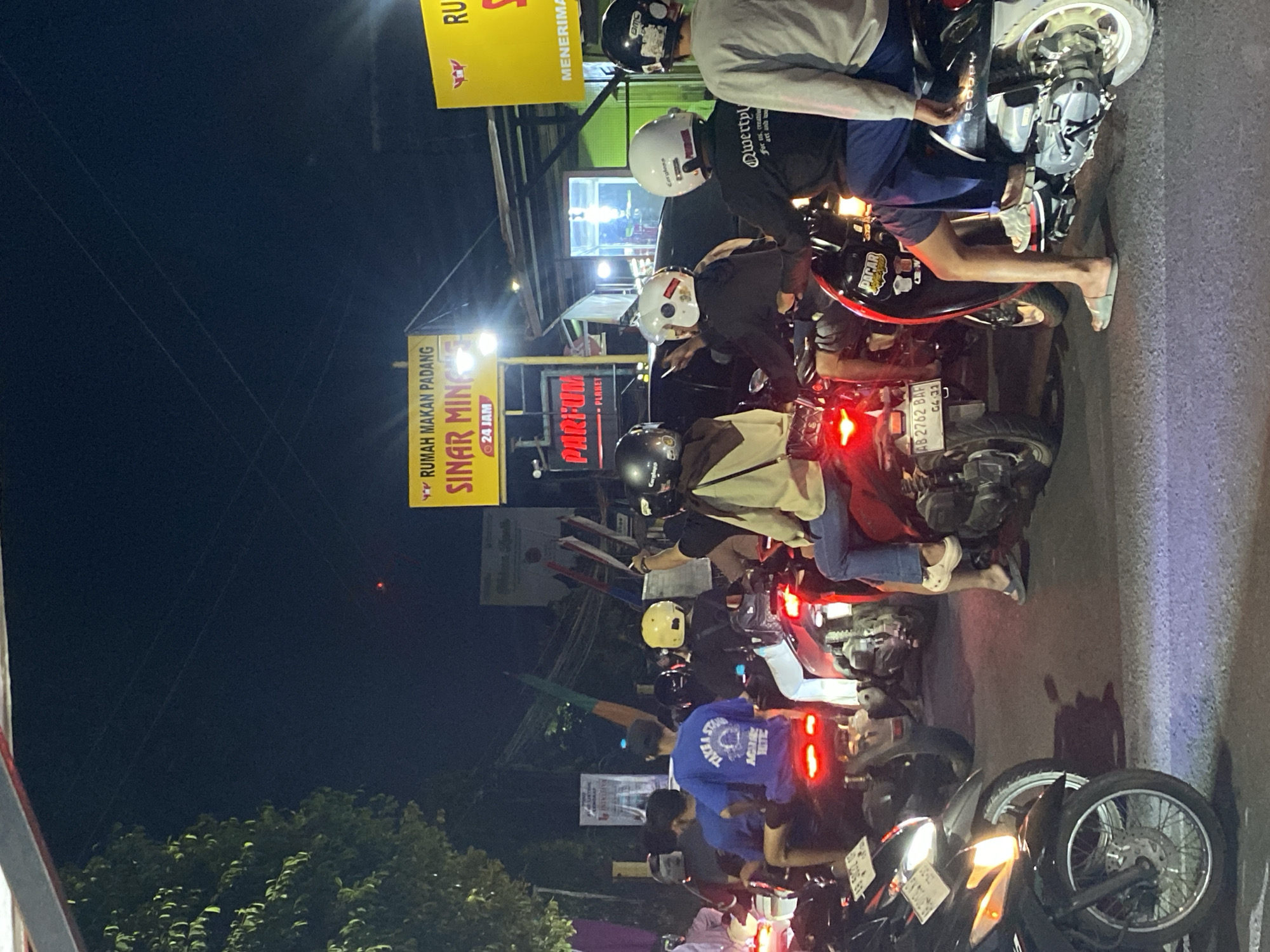 This is a nighttime, eye-level shot of a congested street in Kasihan, Indonesia, likely taken from the perspective of someone on a motorcycle. The scene is dominated by a traffic jam of motorcycles and their riders, illuminated by various artificial light sources. In the background, a yellow neon sign that reads "RUMAH MAKAN PADANG SINAR MING" (Padang Food House Sinar Ming) indicates a restaurant, with "24 JAM" (24 Hours) visible below it. Another yellow sign, partially obscured, appears to say "RUMAH MAKAN" (Food House). A third sign, with red lettering, says "PARFUM PLANET".
The people in the image are mostly young adults, many wearing helmets, sitting on or standing near their motorcycles. Their clothing includes t-shirts, some with graphics like "TAKE A STAND AGAIN'S" and "AGAINST TIME", and jeans or shorts. Some individuals are looking forward, others are interacting with each other or their phones. The motorcycles are parked closely together, some with their headlights and taillights illuminated. The road surface appears to be asphalt with visible tire marks and reflections of lights. The overall atmosphere suggests a busy urban environment at night, possibly a popular gathering spot due to the presence of food establishments and the evident traffic.