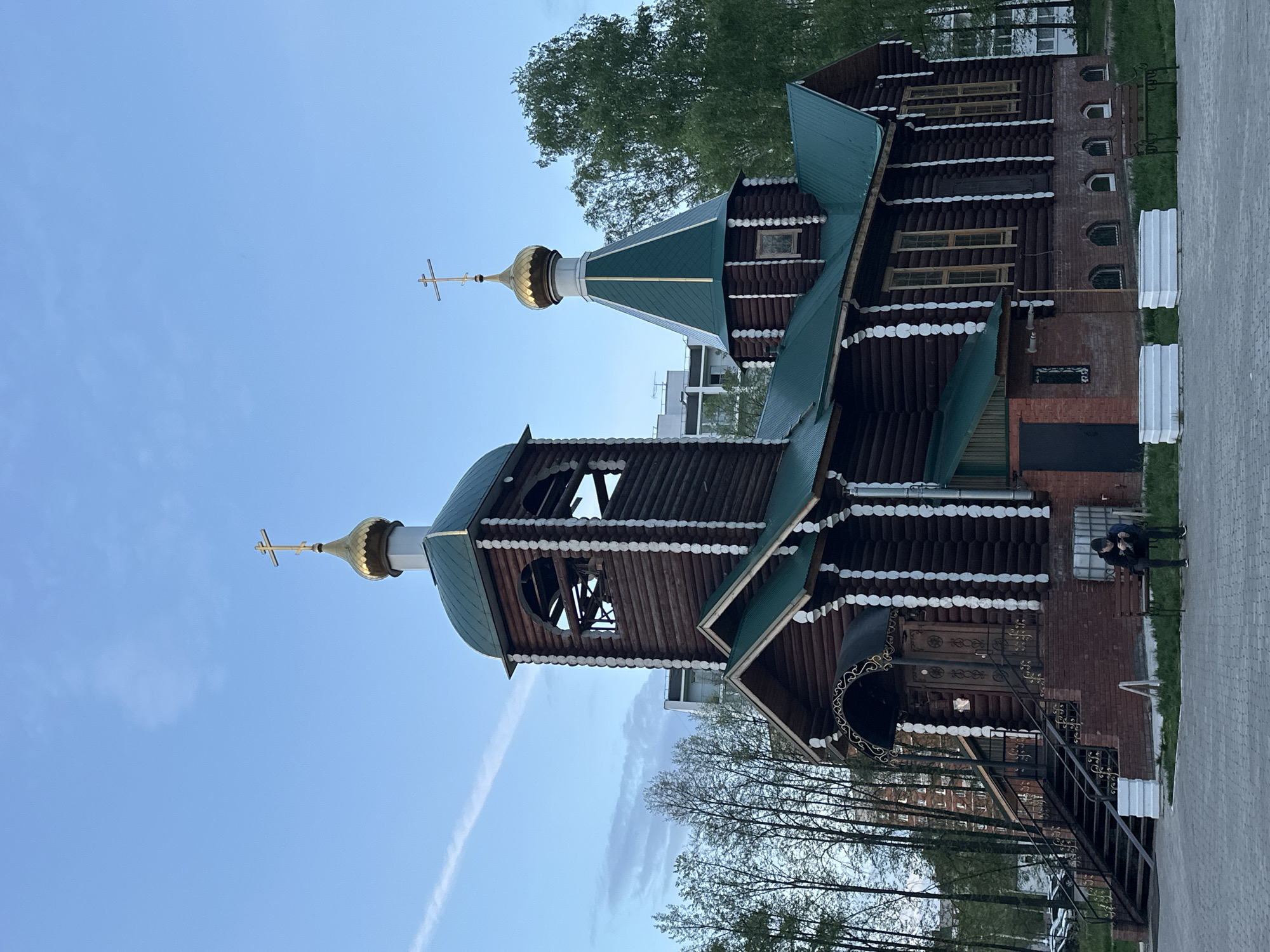 The image shows a wooden Russian Orthodox church in Chaikovsky, Russia. The church has a dark wood exterior with white trim, green roofs, and golden domes topped with crosses. The church appears to be two stories high, with a brick foundation on one side. There are windows visible on both the wooden and brick sections. A wrought iron staircase leads to the entrance. A person sits on a bench next to the building. Paved ground surrounds the building, and trees are visible in the background. The sky is blue with a contrail. Buildings are visible behind the trees in the background.