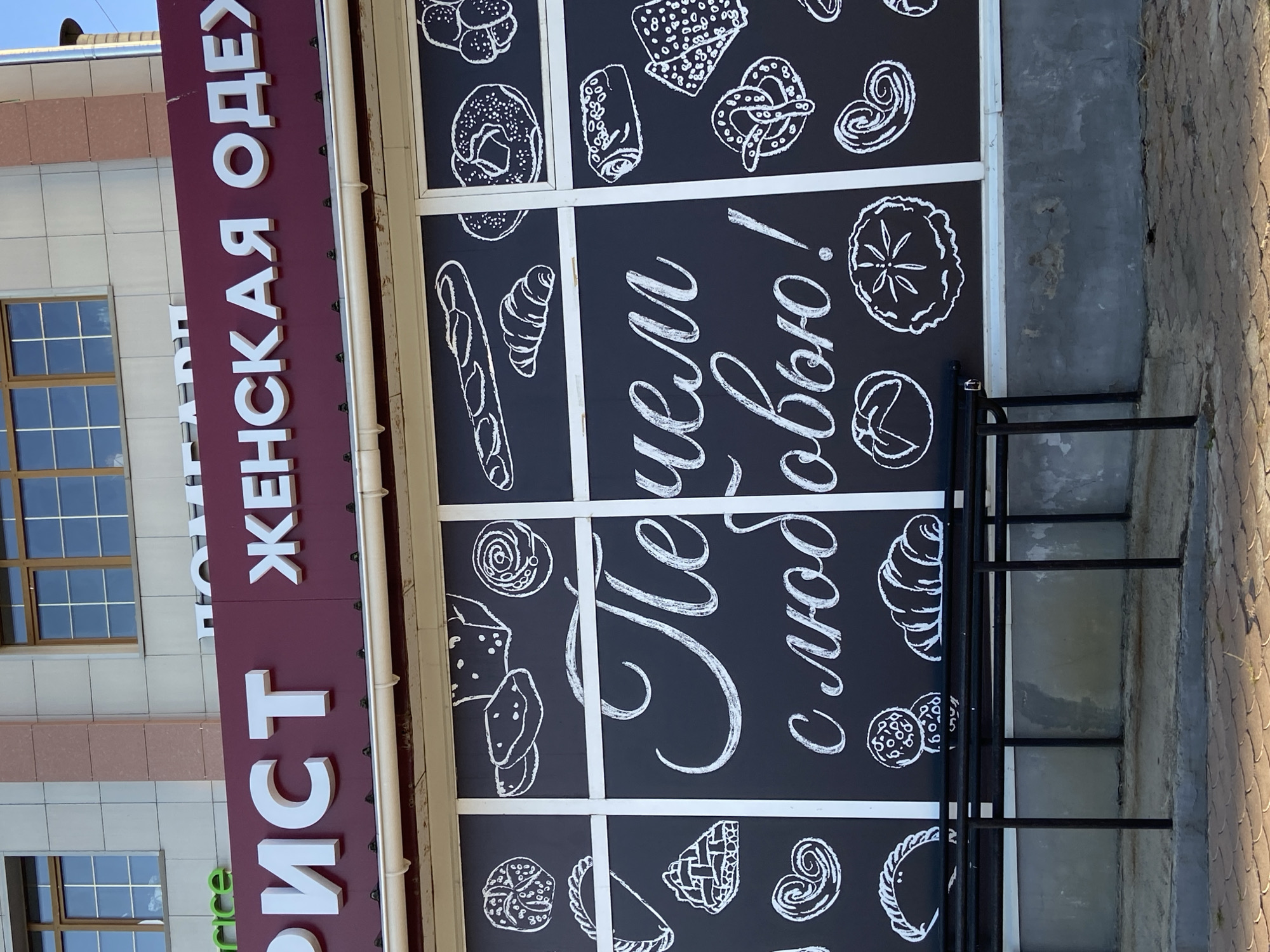 The image shows the storefront of a bakery in Blagoveshchensk, Russia. The store's name, "Печем с любовью!" (Baking with love!), is written in white chalk lettering on a black glass window. Various baked goods, such as bread, croissants, and pretzels, are also depicted in chalk drawings on the window. Above the window, a large red sign displays the words "РИСТ" and "ЖЕНСКАЯ ОДЕЖ" in large white letters, indicating that the building houses both a bakery and a women's clothing store. The building facade features light-colored tiles and windows, suggesting an urban setting. A black metal bench and railing are visible in front of the store.