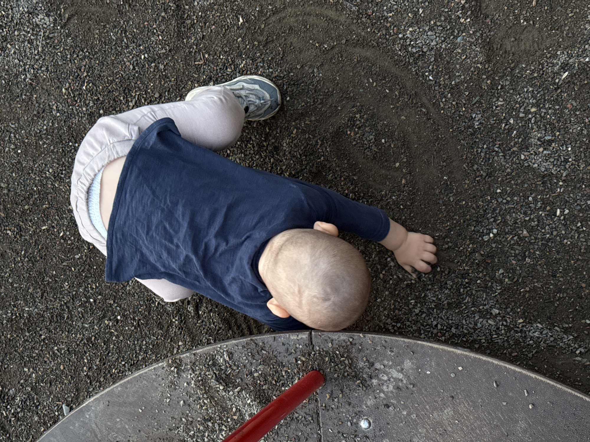 A young child is lying on their stomach on a playground, which appears to be covered in dark gravel and dirt. The child is wearing a navy blue long-sleeved shirt and light grey pants. One shoe, a grey sneaker with blue accents, is visible on their left foot. The child's head is turned to the side, and their right arm is extended with their hand resting on the ground. A red bar and a portion of a textured grey surface, likely part of playground equipment, are in the foreground. The lighting suggests it could be daytime, possibly overcast or late in the day, given the soft shadows. The setting is an outdoor playground, typical of a park or school. No text is visible in the image.