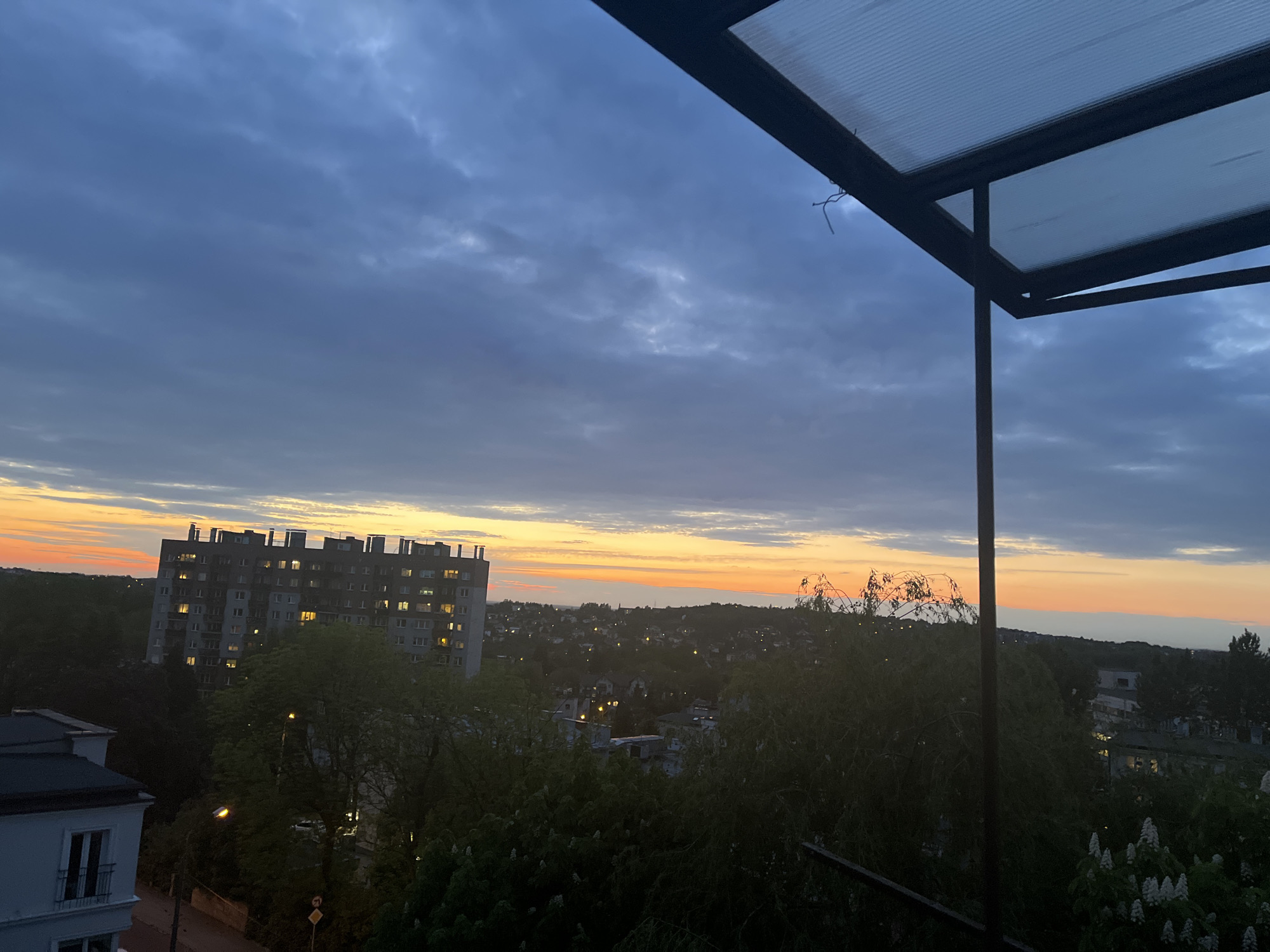 The image depicts a view from a balcony overlooking a city at sunset. A tall apartment building with lit windows is visible on the left, with trees and other buildings filling the space between the balcony and the horizon. The sky is a mix of blue and grey clouds with a strip of orange and yellow light indicating the setting sun. The balcony structure is partially visible in the foreground, framing the scene. The location appears to be an urban area with a mix of residential buildings and green spaces.