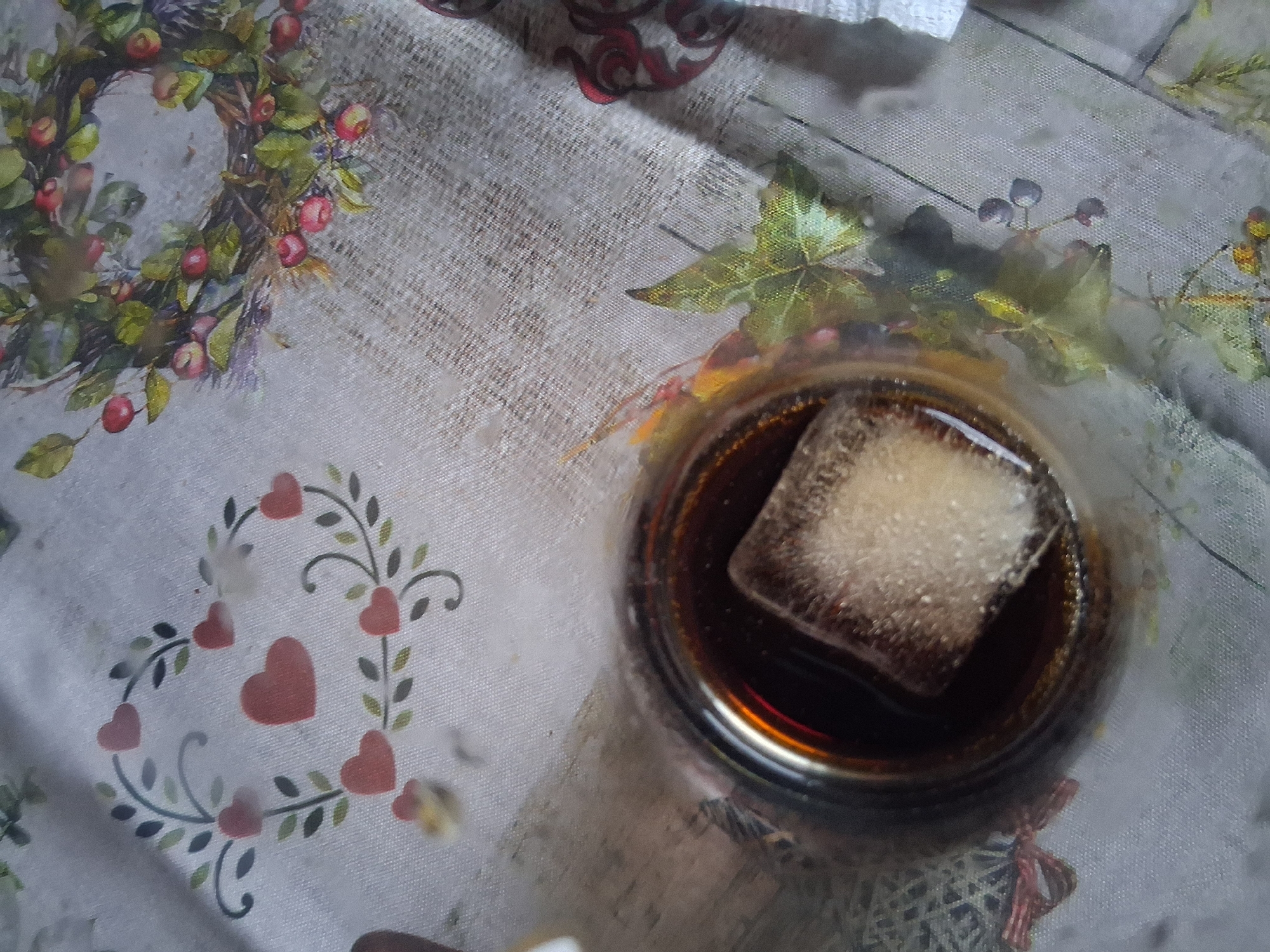 A glass containing a dark liquid and an ice cube sits on a tablecloth. The tablecloth is decorated with a heart-shaped wreath on the left and a circular wreath with berries and leaves on the right. The scene is an indoor setting, possibly a home. The image does not provide any information to indicate a location in Colorno, Italy.