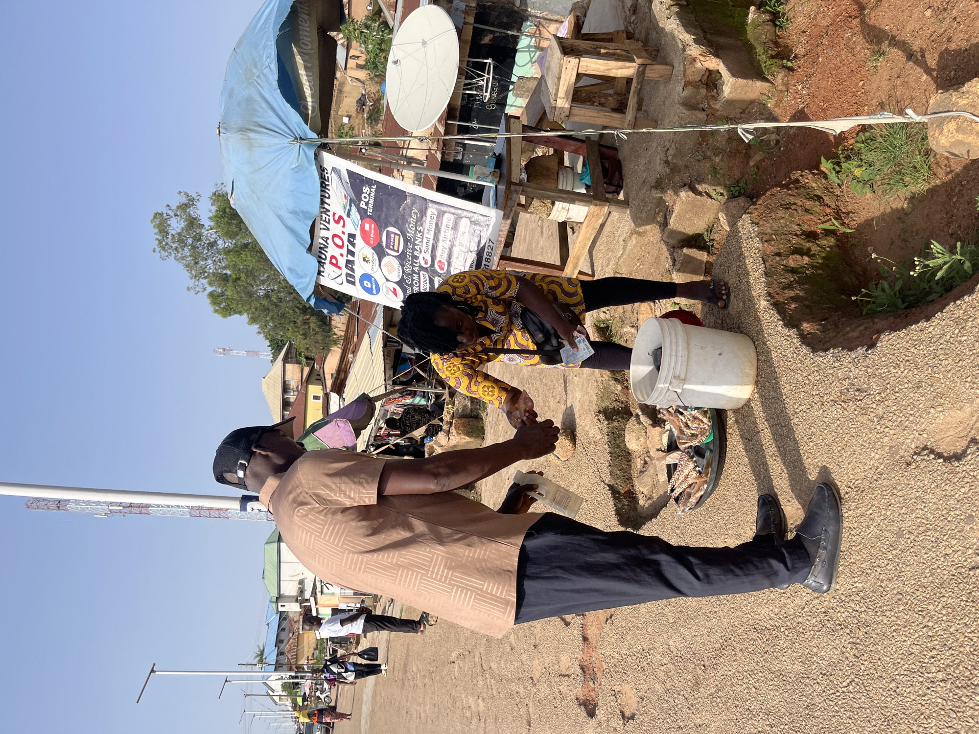 The image depicts a street scene in Jos, Nigeria, with a focus on two individuals engaged in a transaction. A man wearing a brown patterned short-sleeved shirt, dark pants, and a black baseball cap is handing a white bottle to a woman. The woman, dressed in a yellow and black patterned dress and dark leggings, is holding currency. They appear to be at an outdoor stall, indicated by a blue umbrella providing shade over a sign that reads "KAUNA VENTURES & P.O.S DATA". The stall also displays various logos of financial institutions.
The environment is characterized by a dirt road with gravel and debris. In the background, there are several buildings, some with corrugated metal roofs, and a tall telecommunications tower. Other people and vehicles are visible further down the road, suggesting it is a public thoroughfare. A satellite dish is mounted on the side of a structure behind the stall. A white bucket and a tray containing what appear to be dried fish or other food items are also present near the transaction. The ground in the foreground shows a ditch or trench. The lighting suggests it is daytime with clear skies.