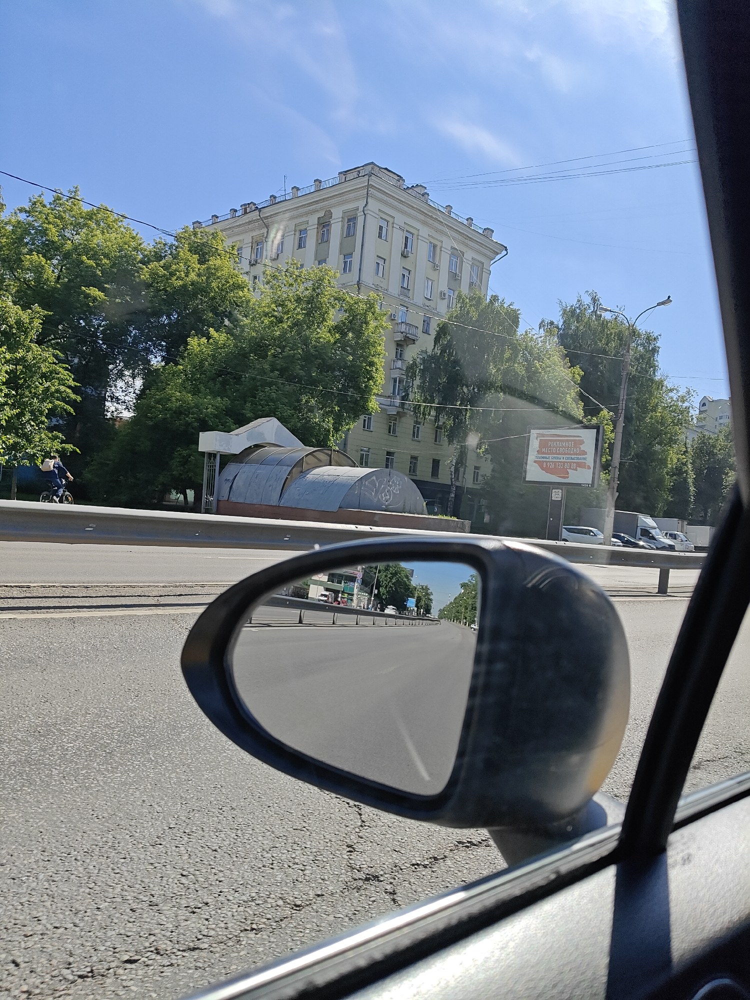 The image captures a view from inside a car in Lyubertsy, Russia, looking out the passenger window and into the side-view mirror. A light-colored, multi-story building with visible air conditioning units is seen in the background, partially obscured by lush green trees. To the left, a person on a bicycle rides along a sidewalk next to a road, wearing a backpack. A bus stop shelter with a distinctive arched, translucent roof is also visible. A billboard with Russian text, which appears to be an advertisement, is mounted on a pole. The side-view mirror reflects a road stretching into the distance, with a clear blue sky and scattered clouds overhead. The road surface is asphalt. The overall scene depicts an urban environment on a bright, sunny day.