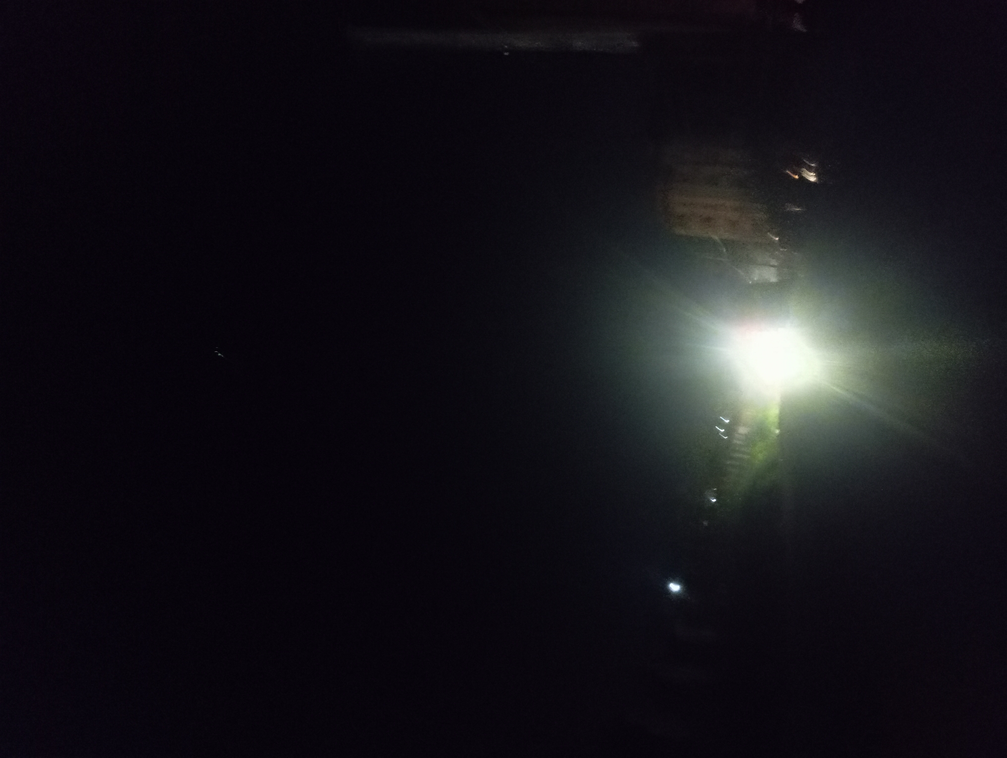 The image appears to be taken at night in Nkpor, Nigeria. A bright light source illuminates a vertical structure, possibly a building or pole, with what appear to be stairs leading up the side. Above the bright light, there's a sign or inscription that's difficult to read. The rest of the scene is dominated by darkness.