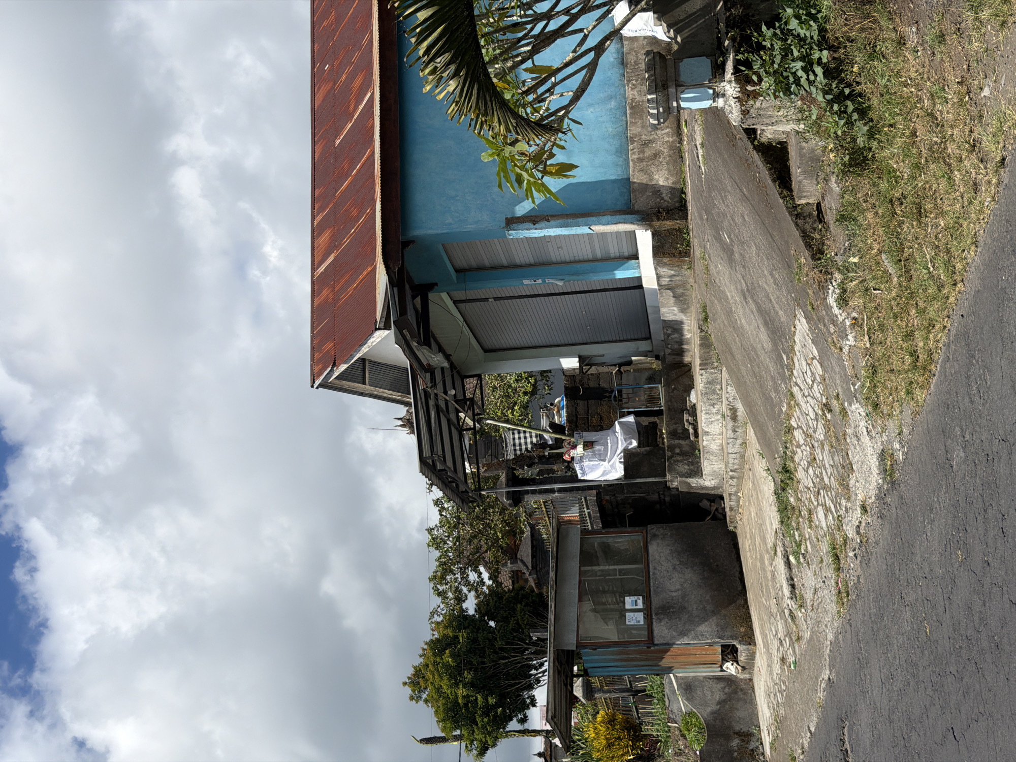 This is an exterior, eye-level shot of a roadside scene in Kintamani, Indonesia, under a cloudy sky. The image is dominated by a bright blue building with a weathered, rusty tin roof. The building has two large, gray roller shutters, suggesting it may be a shop or business. In front of the blue building, a concrete walkway slopes down towards the road. To the left of the blue building, there's a smaller, gray structure with glass panes and a wooden door, possibly a guardhouse or small stall. In the background, lush green trees and some traditional Balinese architecture are visible. A palm frond is present in the foreground on the right side of the image. The road in the foreground is paved and appears aged, with patches of grass and weeds growing through cracks. The overall impression is of a rural or semi-urban commercial area in a tropical climate.
