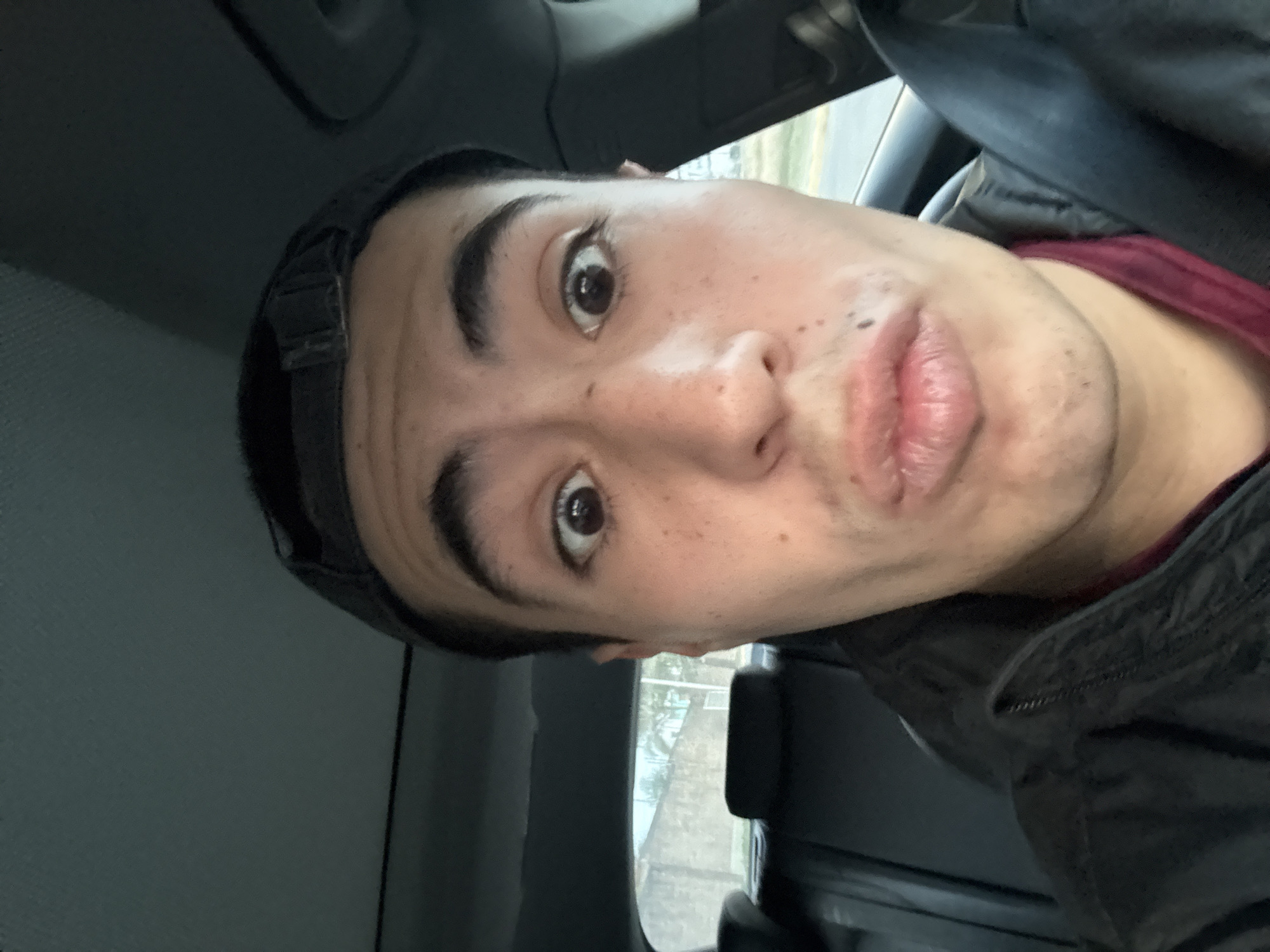 A selfie of a young man inside a car, possibly in San Bernardino Tlaxcalancingo, Mexico. He has dark hair, dark eyebrows, brown eyes, and is wearing a black cap. His lips are pursed in a playful or pouting expression. He is wearing a dark jacket and a burgundy shirt is visible beneath. The car interior is gray. A window is visible behind him.