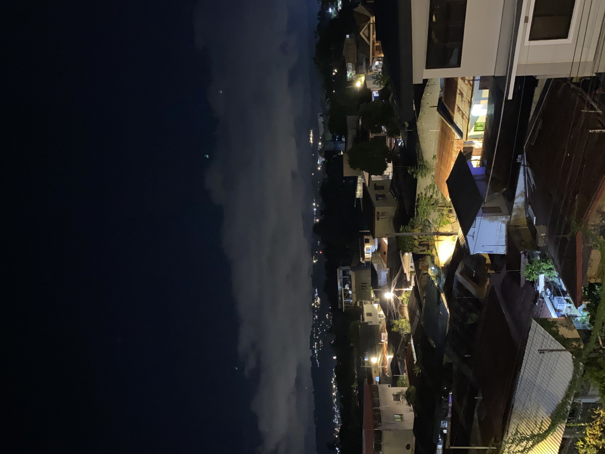 This image presents a nighttime panoramic view of a densely populated urban or semi-urban area within Davao City, Philippines, observed from an elevated vantage point. The dark night sky occupies the upper portion, featuring patches of illuminated cloud cover and several faint, indistinct greenish-bluish light anomalies, possibly atmospheric diffusions or lens flares, near the center.
The mid-ground reveals a range of hills or mountains, their contours faintly visible and dotted with numerous scattered lights indicating distant settlements or infrastructure. The immediate foreground and mid-foreground are dominated by a cluster of closely packed buildings, primarily residential structures with various roof types, including prevalent corrugated metal roofs displaying shades of dark grey, brown, and rust. Artificial lighting emanates from streetlights, illuminating pathways and parts of the buildings, and from within windows and doorways, casting warm glows and pronounced shadows.
Notable objects include utility poles and crisscrossing power lines or communication cables, and various trees and foliage interspersed among the structures, some illuminated from below. On the far right, a section of a multi-story building with light-colored exterior walls and dark windows is visible, suggesting the location from which the photograph was taken. No individuals, direct actions, or interactions are discernible within the frame. The scene is static, defined by the interplay of artificial light against the dark environment.