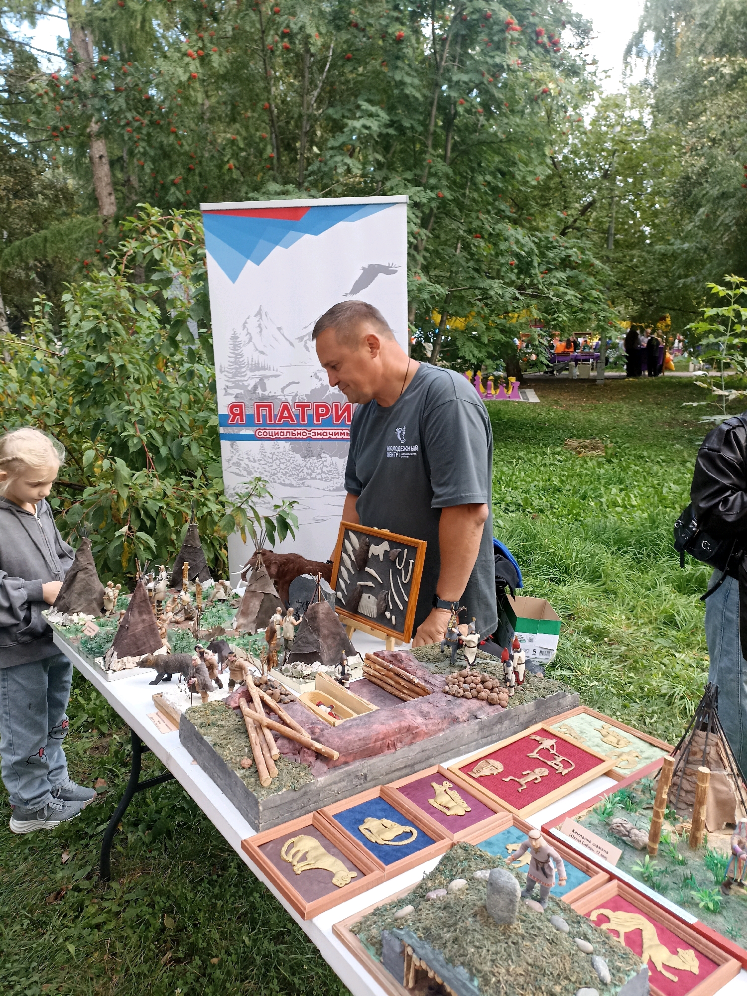 A man and a young girl are standing at a table outdoors, examining a display of miniature dioramas depicting scenes from prehistoric life. The man, wearing a grey t-shirt and a watch, is looking down at the display. The girl, in a grey hoodie and blue jeans, is also looking at the display. The table is covered with various small models, including tipis, figurines of people and animals, and depictions of primitive tools and hunting scenes. In the background, a large banner with Russian text and imagery of mountains and a bird is visible. Trees and grass surround the scene, suggesting a park or outdoor event. The city of Novosibirsk, Russia, is implied as the location based on the provided context.