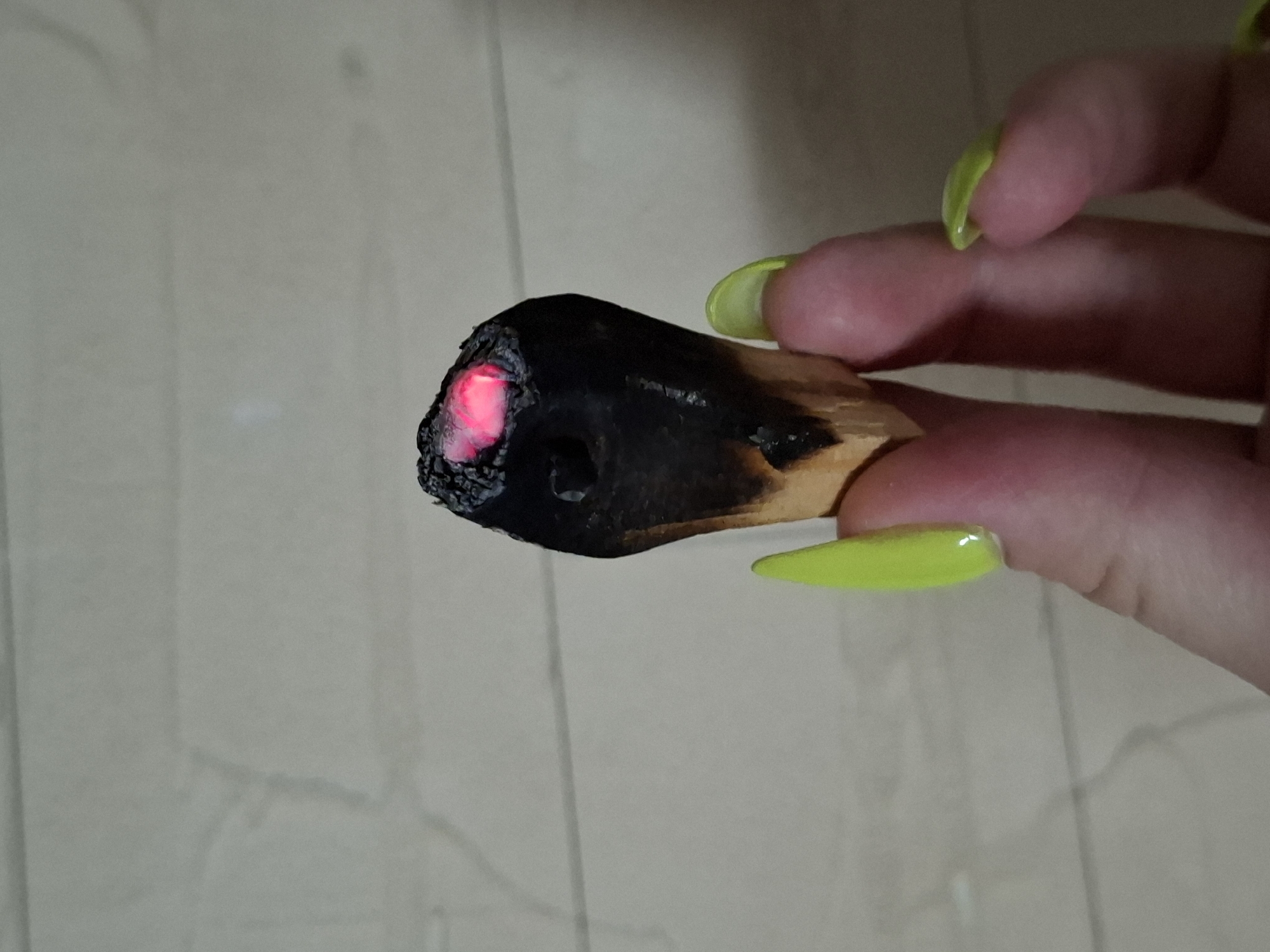 A close-up photograph shows a person's hand holding a piece of charred wood that is glowing red at one end. The fingers of the hand have long, neon yellow painted nails. The background appears to be a plain, light-colored surface. The location is not discernible from the image.