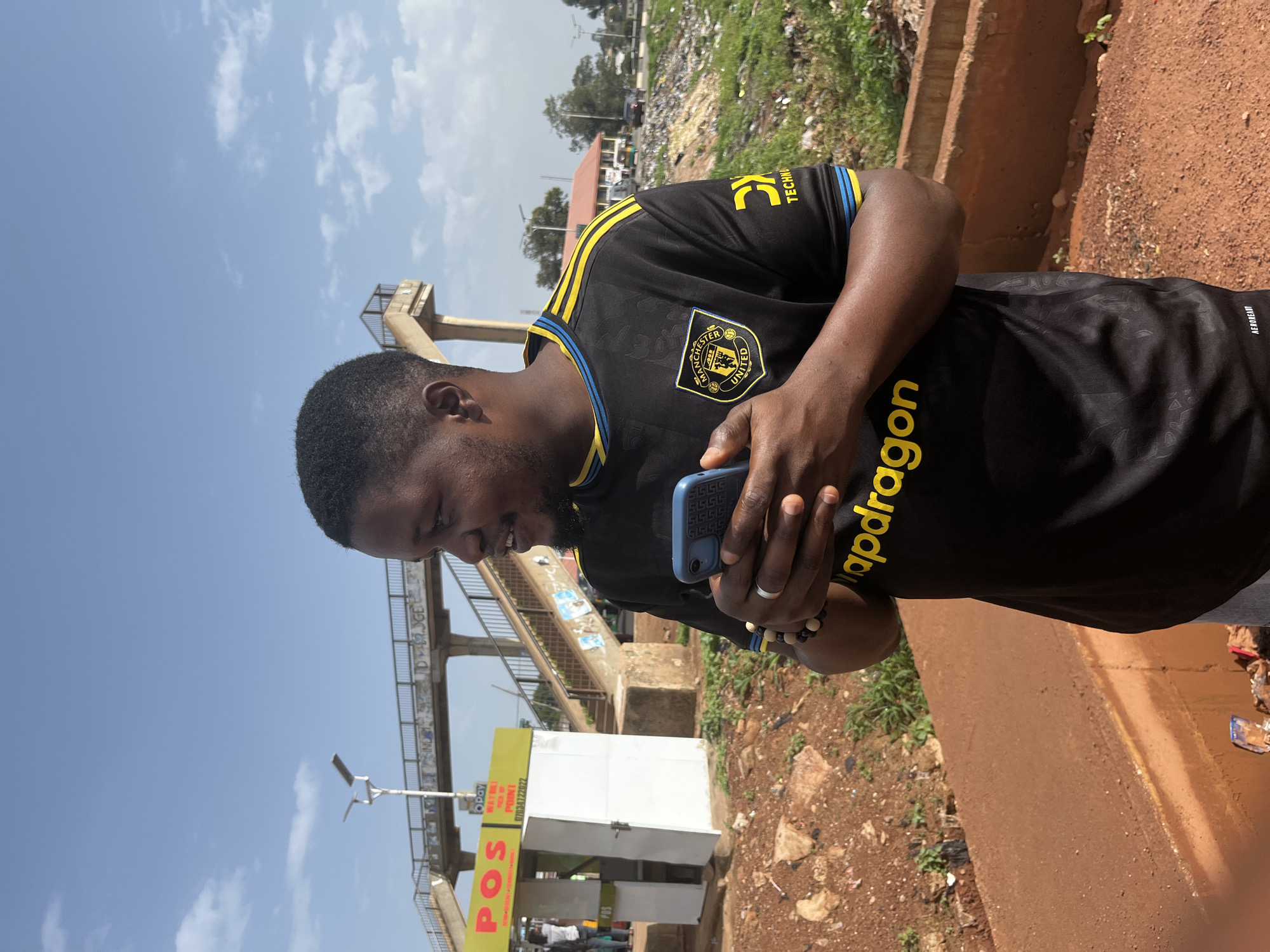 A man stands outdoors in Gura Top, Nigeria, holding a blue smartphone in his hands. He is wearing a black Manchester United football jersey with yellow and blue accents. In the background, a pedestrian bridge crosses over a road. To the left of the bridge, a white structure with yellow signage that reads "POS" is visible. The ground in the foreground is reddish-brown earth with scattered rocks and vegetation. The sky is blue with scattered white clouds.