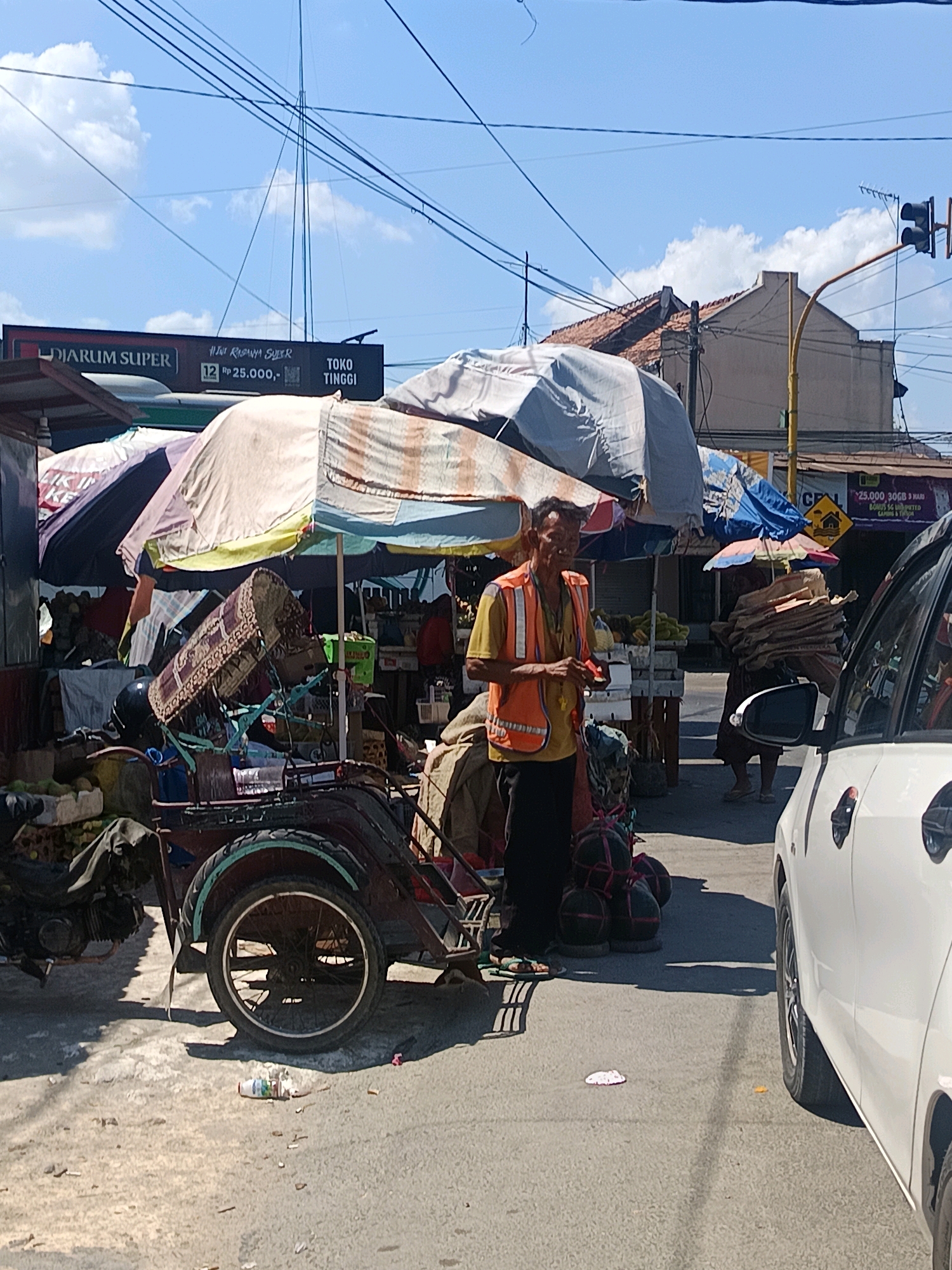 The image captures a street scene in Blega, Indonesia, on a sunny day. In the foreground, a white car is parked on the right side of the road. To the left of the car, a man wearing a yellow shirt and an orange safety vest stands near a three-wheeled vehicle, possibly a becak or a modified motorcycle. The vehicle is laden with various items, including what appears to be a large, patterned textile covering a cylindrical object, and several large, round bundles wrapped in dark material with red string. Behind the man and the vehicle are several market stalls covered by awnings, displaying goods. A sign reading "DIARUM SUPER" and "TOKO TINGGI" is visible in the background. Further down the street, a traffic light is visible, and another person is seen carrying a stack of cardboard. The ground is paved with asphalt and shows scattered debris. Power lines crisscross the sky, and a few clouds are present in the blue sky.