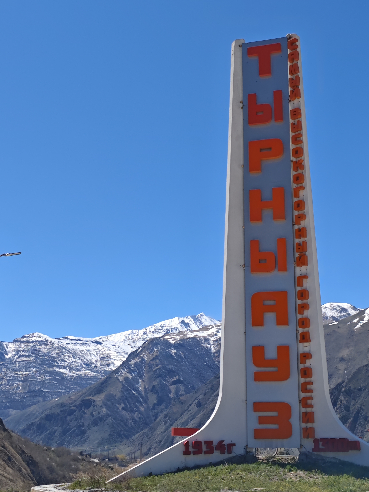 The image shows a tall sign marking the location of Tyrnyauz, Russia. The sign is primarily white with large, orange Cyrillic letters spelling out "Tyrnyauz". To the right of these letters, also in orange, are additional Cyrillic words. The sign has a stylized design with wide bases that taper inward as it rises. At the bottom left of the sign, in red, is the year "1934г". On the bottom right is "1300м" written in red.
Behind the sign is a mountainous landscape with snow-capped peaks. The sky above is a clear, bright blue. The base of the sign sits on a small patch of green vegetation. The lighting suggests it is a sunny day. A street lamp is visible on the far left of the image.