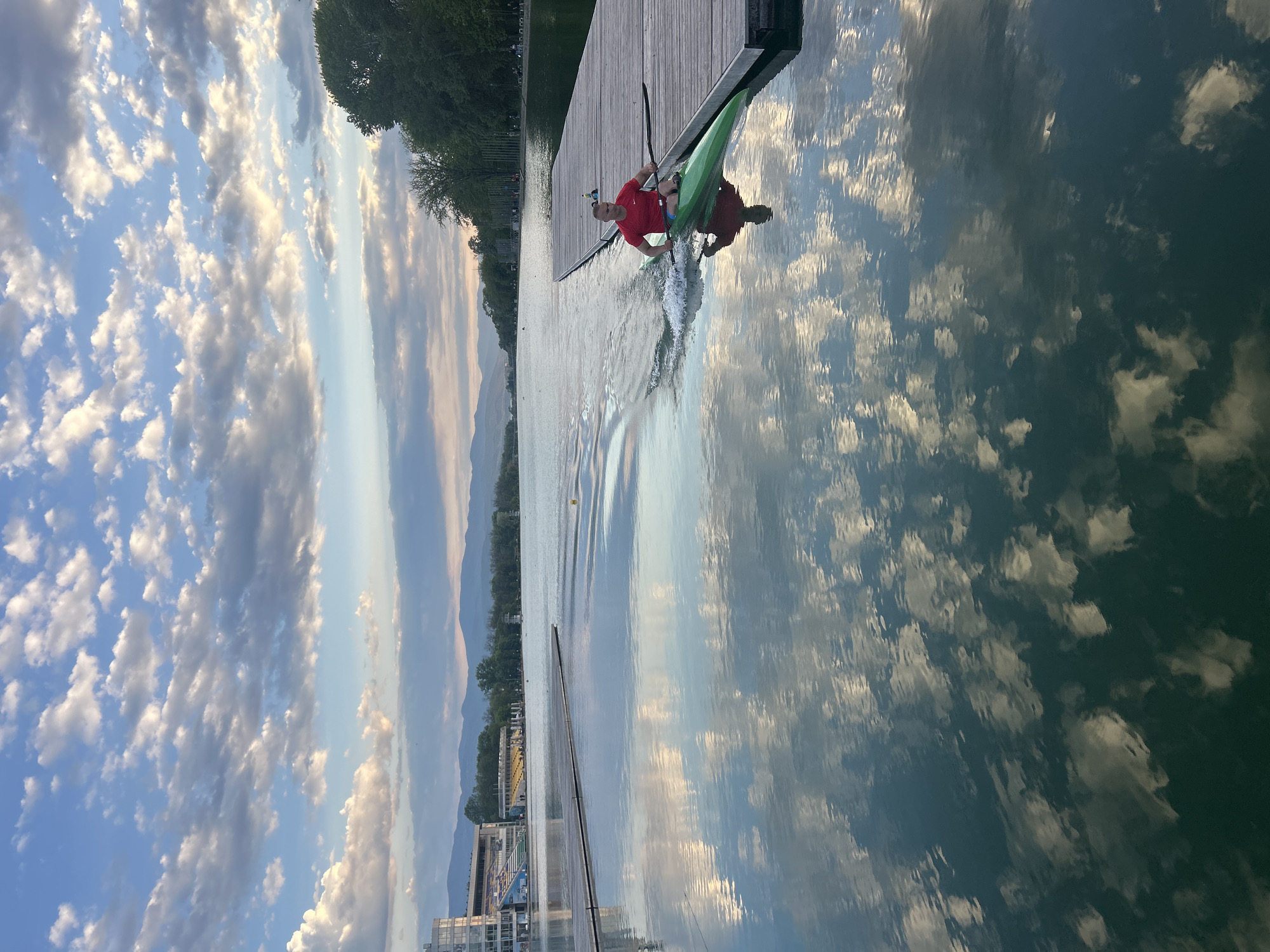 A landscape-oriented photograph shows a man in a kayak on the Plovdiv Rowing Canal in Plovdiv, Bulgaria. The man, wearing a red shirt, is positioned on a green kayak close to a wooden dock on the right of the image. He is holding a paddle and creating a wake in the water. The canal reflects the cloudy sky above. In the background, across the canal, are trees, buildings, and distant mountains. Part of the city's infrastructure is visible on the left of the scene. The sky is filled with white and grey clouds, while the water mirrors the sky's color.