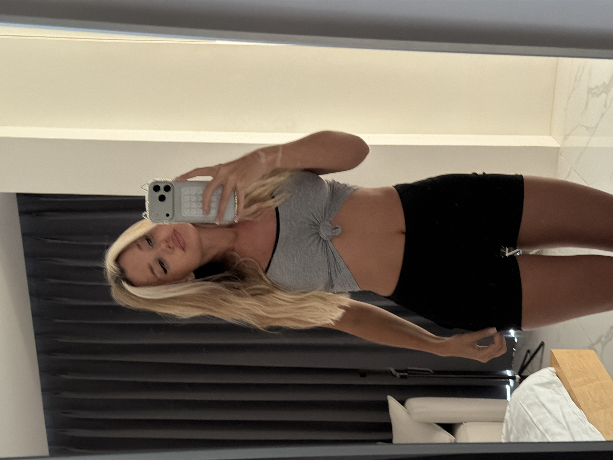A young woman with blonde hair tied in a high ponytail takes a mirror selfie in what appears to be a bedroom. She is wearing a grey tied-up crop top and black shorts. Her phone, with a light-colored case, is held up to take the photo. In the background, a grey curtain covers a window, and to the left, a white headboard of a bed is visible. The floor is tiled. The overall scene is a casual, indoor setting.