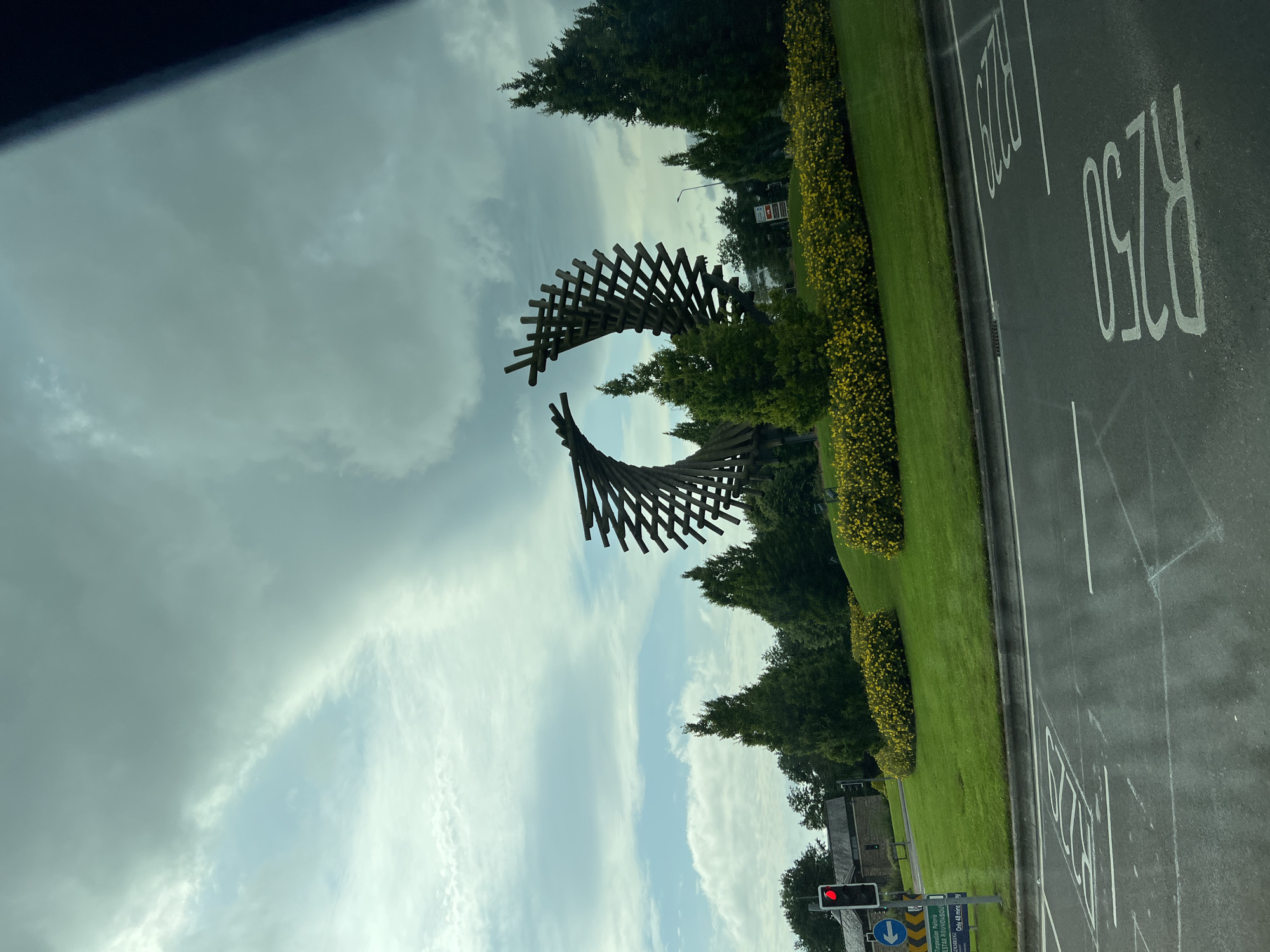 A modern wooden sculpture, resembling two intertwined circular forms, stands in the center of a roundabout in Letterkenny, Ireland. The sculpture is composed of numerous angled wooden beams, creating a striking geometric pattern against the cloudy sky. The roundabout itself is covered in lush green grass and lined with vibrant yellow flowering bushes. To the left of the roundabout, a traffic light displays a red signal, and a blue directional sign points to "Letterkenny Roundabout." Behind the roundabout, a building with a brick facade is visible, along with several evergreen trees. The foreground shows a section of asphalt road with white road markings, including lane indicators. The overall scene suggests a well-maintained public space on a somewhat overcast day.