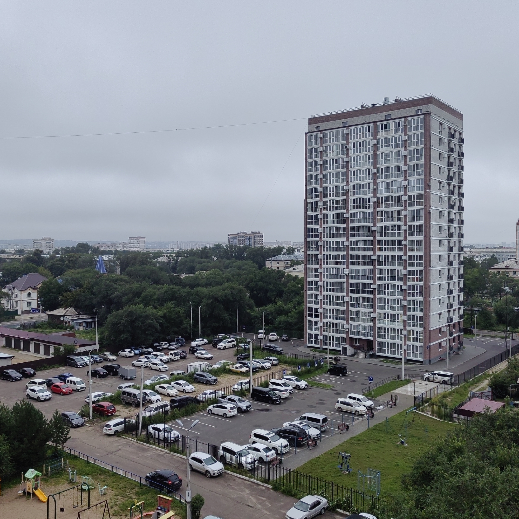 The image presents an aerial view of a residential area in Blagoveshchensk, Russia, under an overcast sky. Dominating the right side of the frame is a tall, multi-story apartment building with a light-colored facade and numerous windows, punctuated by reddish-brown vertical accents. A parking lot with numerous cars is situated in front of the building. Beyond this, a dense area of green trees separates the apartment complex from other, lower-rise buildings and a distant cityscape. A children's playground with swings and slides is visible in the lower left quadrant, adjacent to a grassy area and a fence. In the foreground, more vehicles are parked in a lot. The scene depicts a typical urban environment with residential buildings, infrastructure, and natural elements. There are no people or active interactions visible in the image.