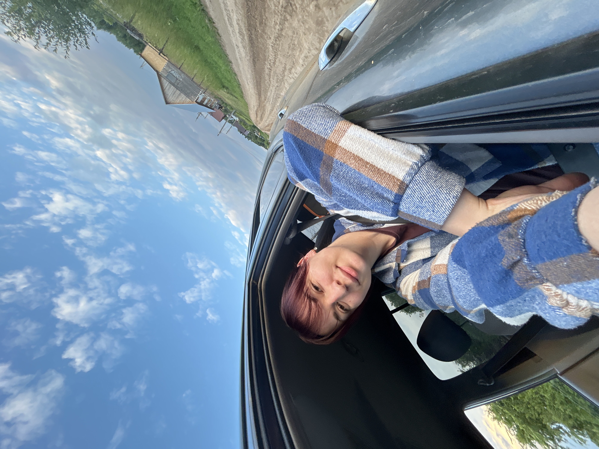 In a selfie taken from inside a vehicle, a person with short, reddish-brown hair leans out of the open passenger window. They are wearing a blue and brown plaid shirt and are looking at the camera with a slight smile. The vehicle appears to be on a dirt road, with a rural landscape visible outside. The road curves ahead, leading towards a few wooden buildings in the distance. The sky is a clear blue with scattered white clouds. The overall scene suggests a leisurely drive through a rural area in the vicinity of Фоки, Russia.