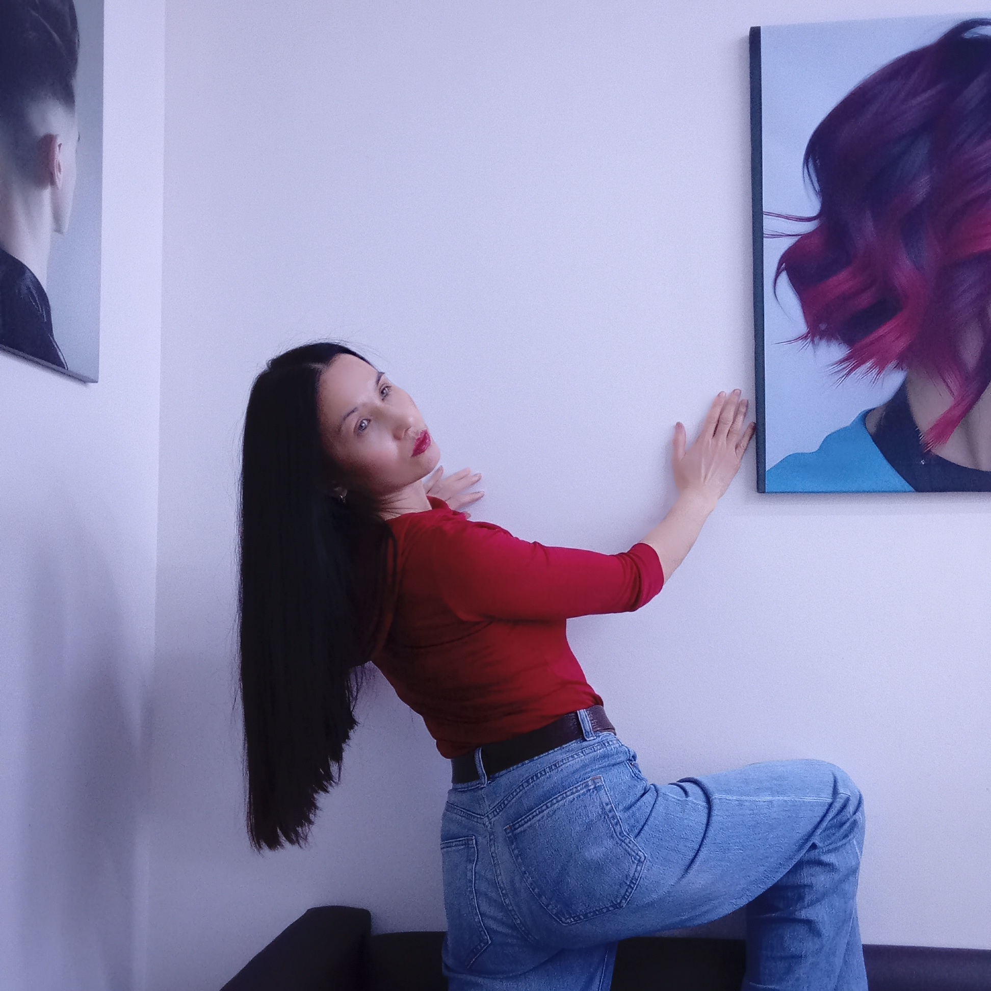 A woman with long, straight dark hair and fair skin is posing indoors in Izhevsk, Russia. She wears a long-sleeved red top, high-waisted blue jeans with a dark belt, and red lipstick. The woman leans against a plain, light-colored wall, with her right knee bent and resting on a dark couch or bench, partially visible at the bottom of the frame. Her left hand is pressed against the wall, while her right hand is near a framed artwork. She gazes upwards and slightly to her right, displaying a composed expression.
Two canvas prints are visible on the wall. The one on the left shows the back of a person's head with a short, stylized haircut. The print on the right features a close-up of a person with vibrant, wavy reddish-pink hair and a light blue garment. The interior space has light, possibly pale purple, walls and soft, even lighting, lending a cool tone to the scene. The setting appears to be a minimalist room, possibly a salon or art display area, suggested by the hair-themed artworks. No visible text is present.