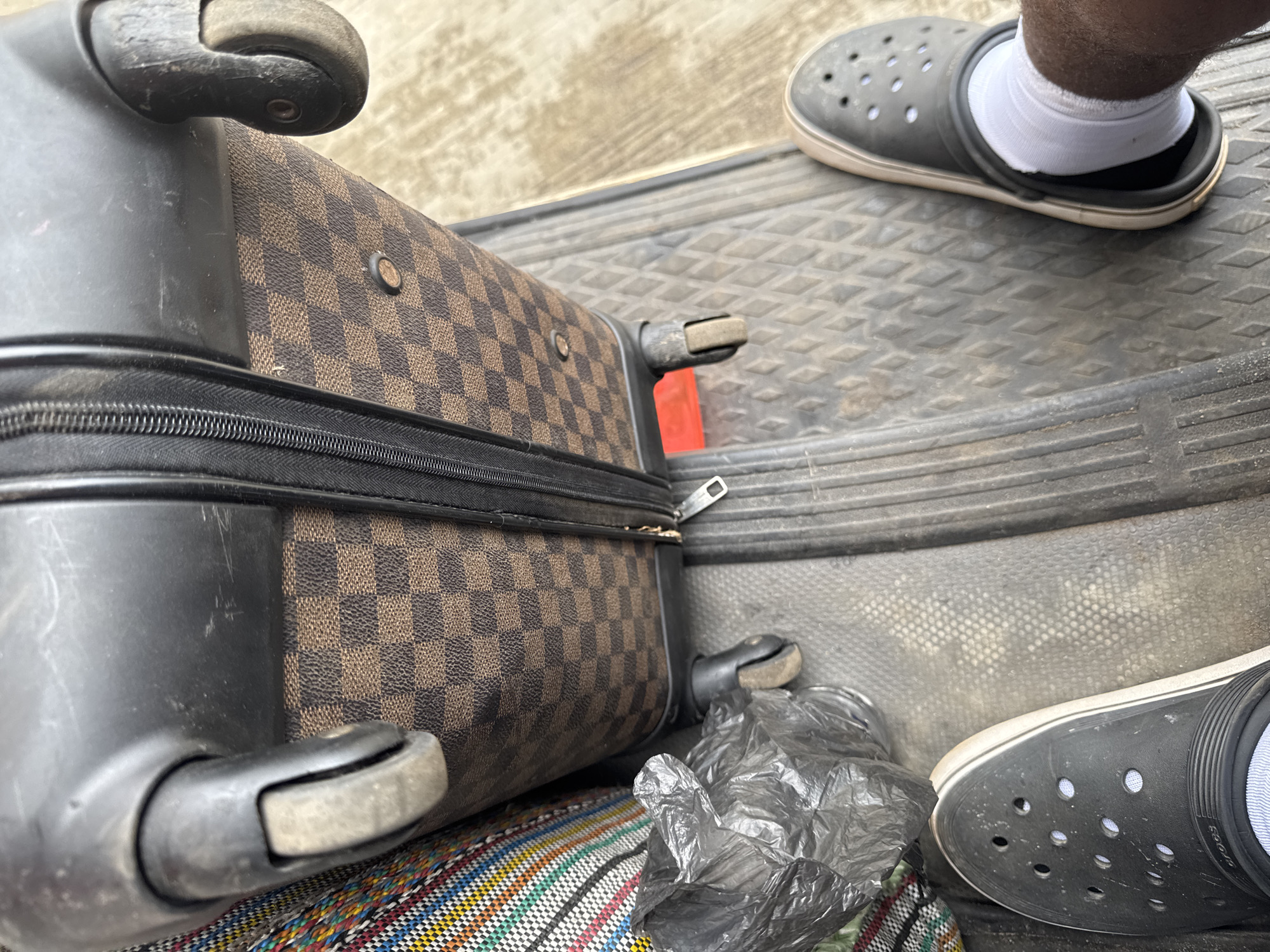 The image shows a brown and black checkered rolling suitcase, partially visible, with its wheels and a zipper. The suitcase sits on a multicolored striped cloth and a crumpled gray plastic bag. Two feet wearing gray Crocs with white socks are also in the frame. The scene is set on a gray textured surface. The location is Ijanikin, Nigeria.