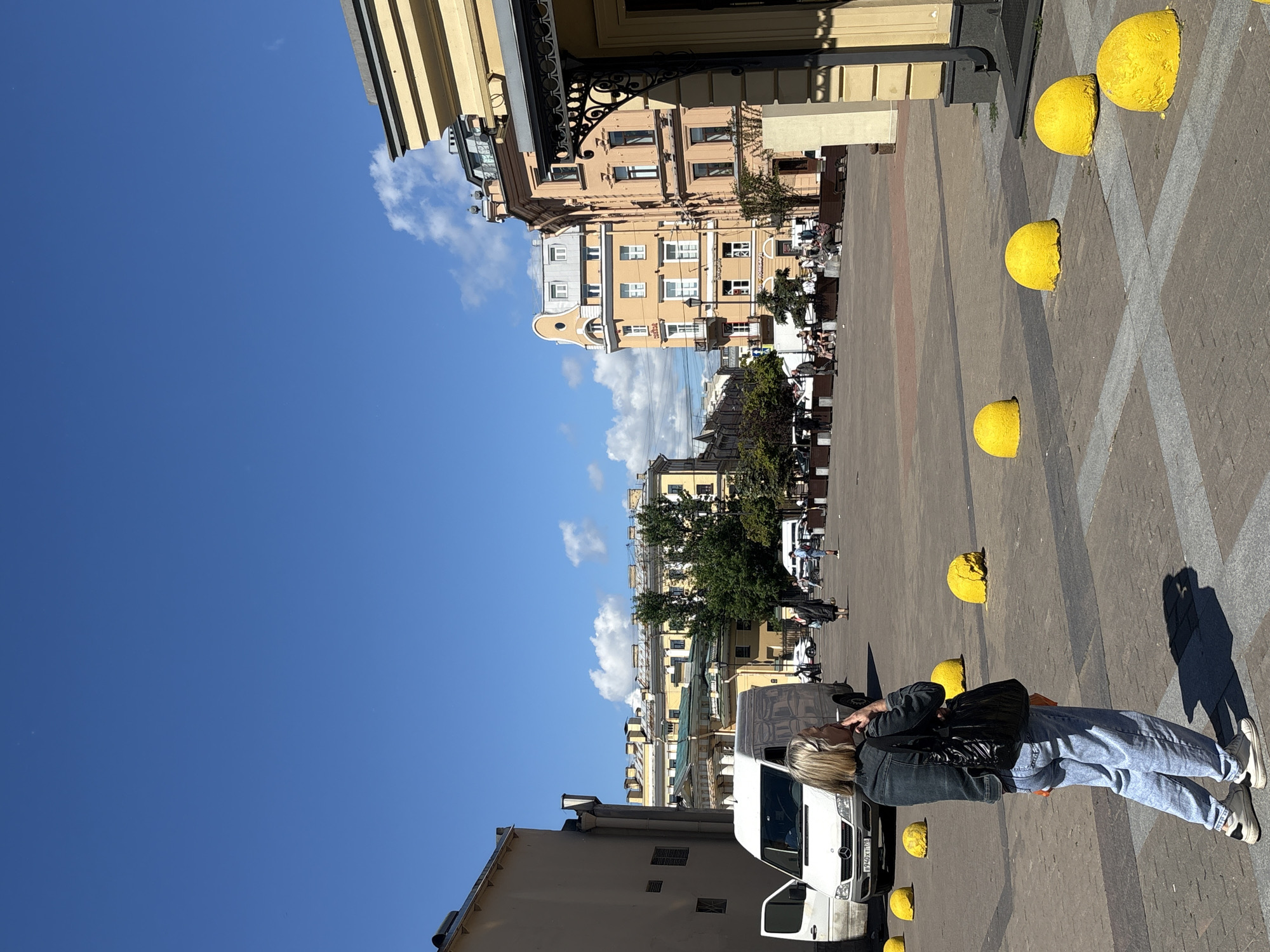 A woman with blonde hair is standing in a sunlit outdoor plaza in Saint Petersburg, Russia. She is wearing a denim jacket, light blue jeans, and white sneakers, and she is holding a black bag over her shoulder. The plaza is paved with cobblestones and lined with several bright yellow spherical bollards. In the background, there are buildings with light yellow facades, trees, and a clear blue sky with scattered clouds. A white van is parked to the left of the woman, and other people and vehicles are visible in the distance. The scene appears to be a public square, possibly in a historic part of the city, given the architecture.