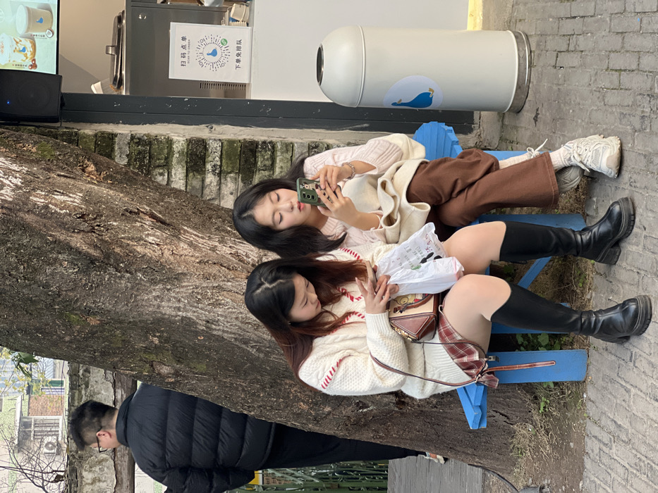 The image depicts an outdoor urban scene in Yuzhong District, China, under daylight conditions. Two East Asian females are seated side-by-side on a blue slatted wooden bench. The female on the left wears a white cable-knit sweater with red stripe accents, a red plaid mini-skirt, and black platform knee-high boots. She holds a brown leather cross-body bag and a translucent white plastic shopping bag, looking down at a smartphone in her hands. The female on the right wears a light pink ribbed sweater, brown wide-leg trousers, white ankle socks, and white athletic shoes. She holds a green-cased smartphone with a finger ring holder, actively viewing its screen.
They are positioned in front of a large tree trunk. To their right, a segment of a light-colored brick wall is visible, with a black speaker mounted above eye level. Adjacent to the bench on the right is a white vendor counter with an overhead digital display showing beverage images. A white sign on the counter displays Chinese characters "扫码点单" (Scan to Order) and "下单免排队" (Order without queuing), along with a blue duck logo and a QR code. A white cylindrical trash receptacle, also featuring a blue duck logo, stands immediately to the right of the bench. In the upper left background, an East Asian male in a black puffer jacket stands with his back partially to the viewer, near a wall. Beyond him, a building with an external air conditioning unit is visible. The ground is paved with rectangular stone bricks.