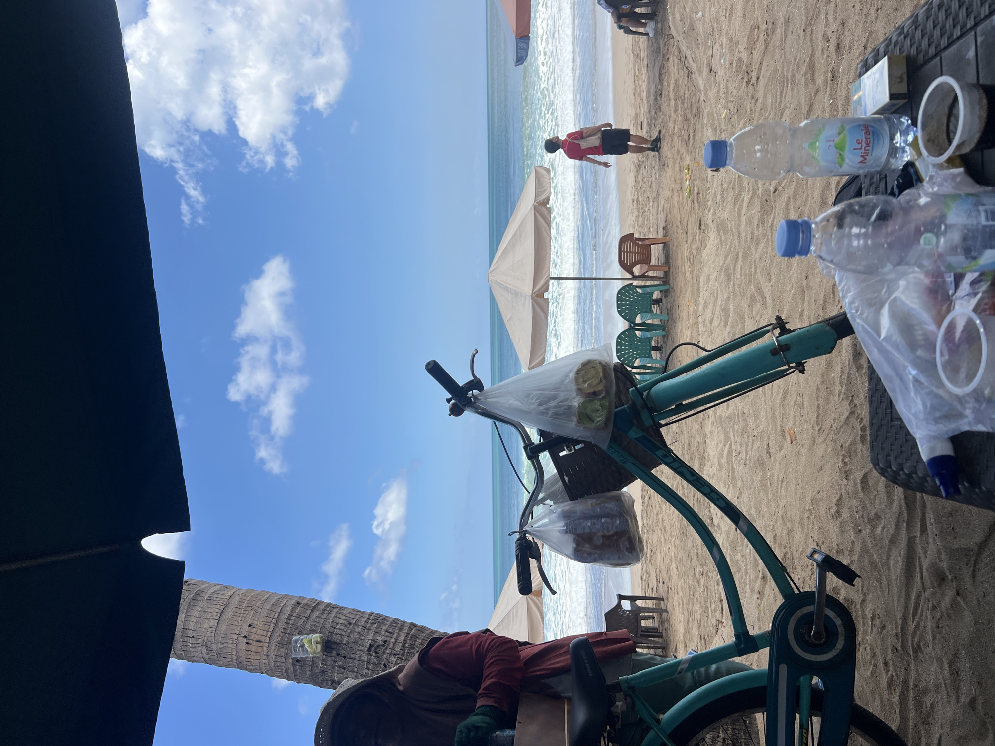 A beachfront scene in Kuta, Indonesia, features a turquoise bicycle parked on the sand. The bicycle has a basket and bags hanging from its handlebars, suggesting it is used for selling goods. In the background, the ocean stretches to the horizon under a blue sky with scattered clouds. Several beach umbrellas and chairs are set up on the sand, and a few people are visible, including one walking near the water. The foreground shows a table with plastic water bottles, a cigarette pack, and a cup of coffee. The overall atmosphere is one of a relaxed beach setting, possibly a vendor's stall.