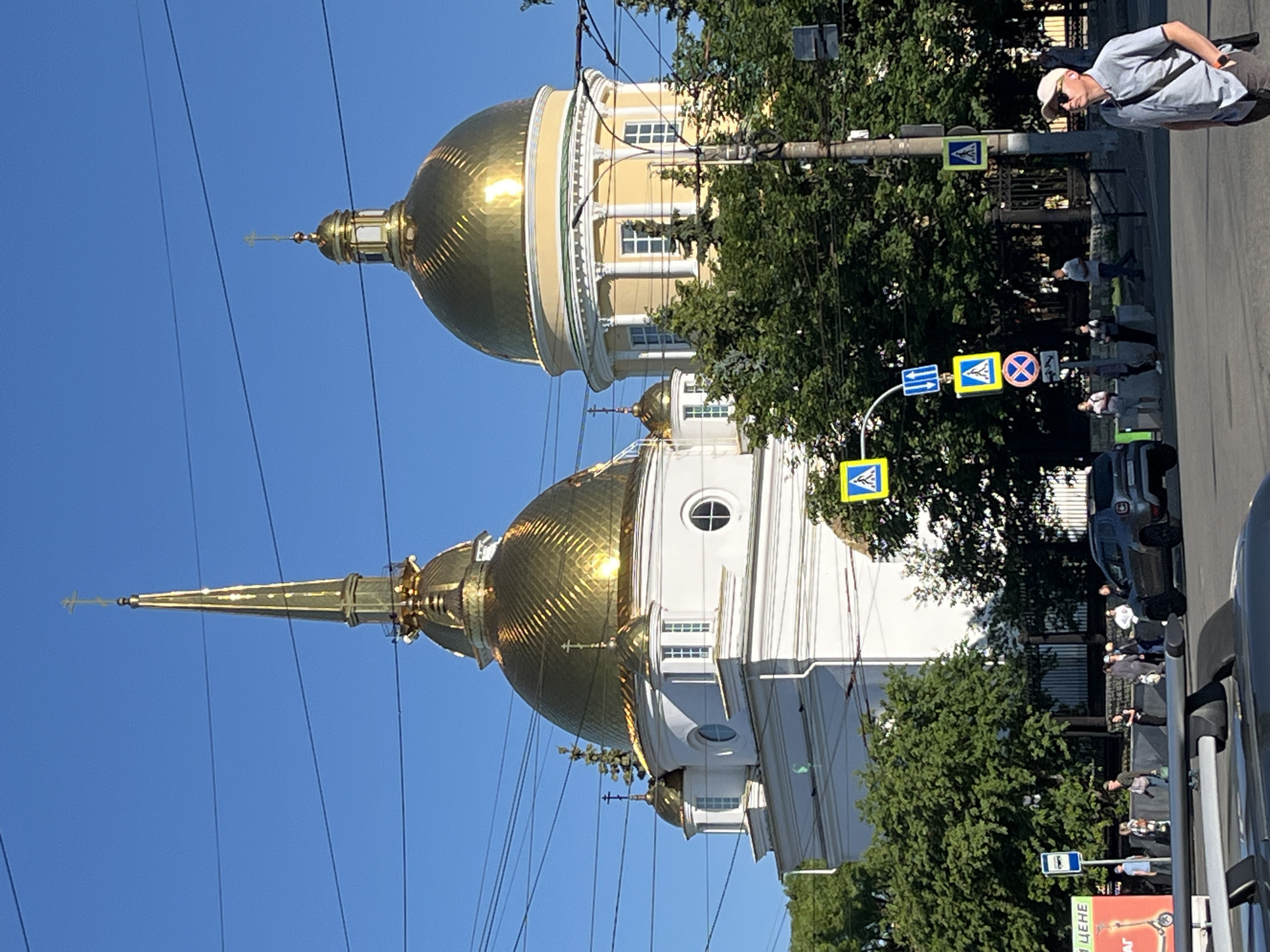 The image depicts the Holy Trinity Cathedral in Izhevsk, Russia, on a clear day with a blue sky. The cathedral features prominent golden domes, one being a tall spire and the other a large, rounded dome. The building is white with architectural details. In the foreground, there are various road signs, including pedestrian crossing signs and a bus stop sign. A few people are visible on the sidewalk and crossing the street, some walking, some standing. A dark gray SUV is parked near the right side of the image. Trees line the street, and power lines crisscross the sky. A man wearing a light-colored cap and sunglasses is standing on the right side of the street, looking upwards.