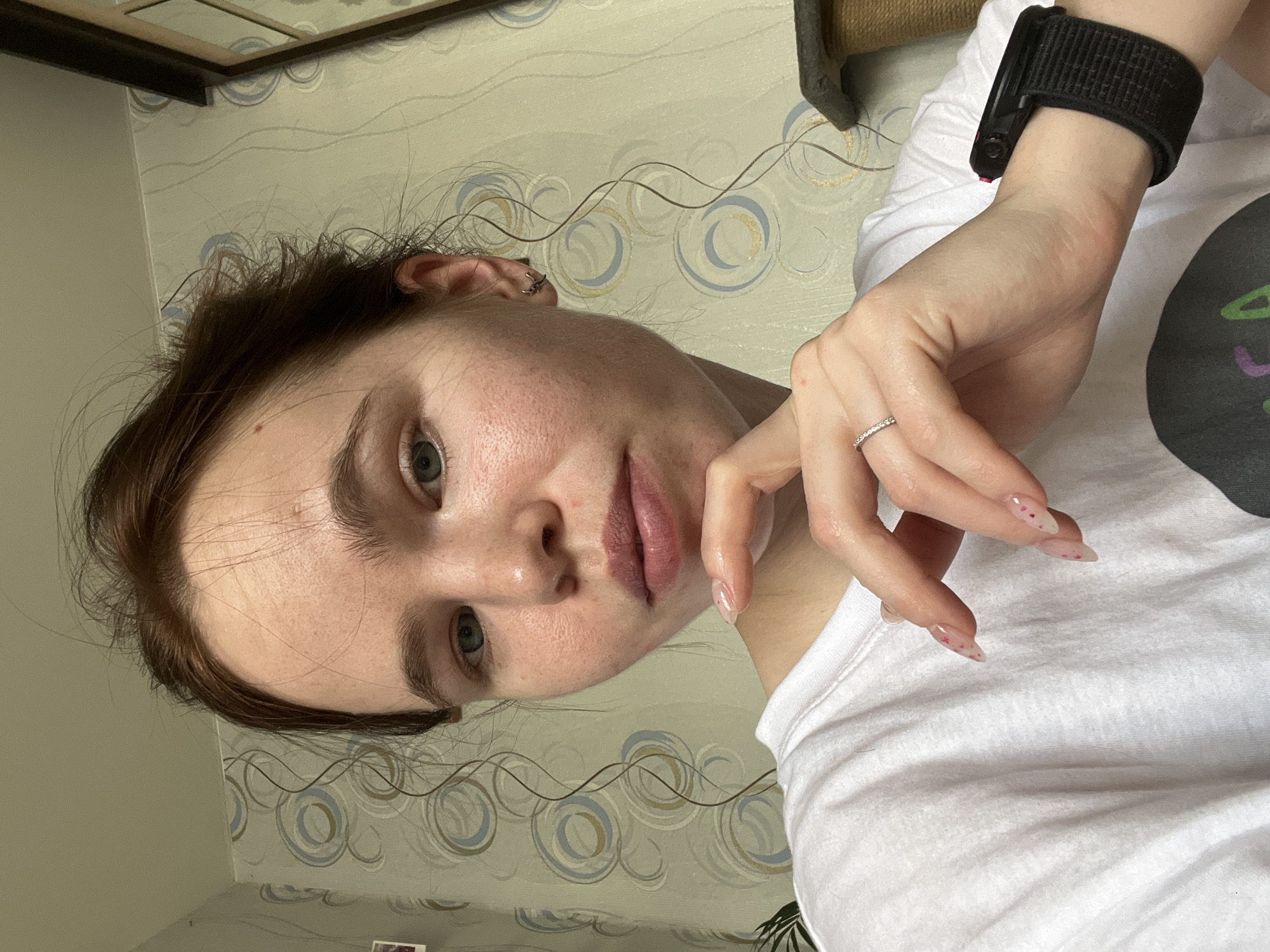 A young woman is captured in a vertical selfie, holding her left hand near her chin while looking directly at the camera with a neutral expression. She has light skin, dark brown hair, and light-colored eyes. She is wearing a simple white short-sleeved t-shirt.
Notable accessories include a black smartwatch or fitness tracker with a fabric strap on her right wrist, and a thin, silver-colored ring with small stones on the ring finger of her left hand. Her fingernails are long, pointed, and translucent, adorned with small pink or red speckles. A small ear piercing is visible on her left ear.
The background reveals an indoor setting, likely a home in Chaikovsky, Russia, with patterned wallpaper featuring light blue, grey, and brown circular and wavy motifs. A dark wooden-framed mirror or picture frame is partially visible in the top left corner, and a textured, possibly fabric-covered item, resembling a scratching post for a pet, is seen in the upper right. The lighting suggests it is daytime. There is no visible text in the image.